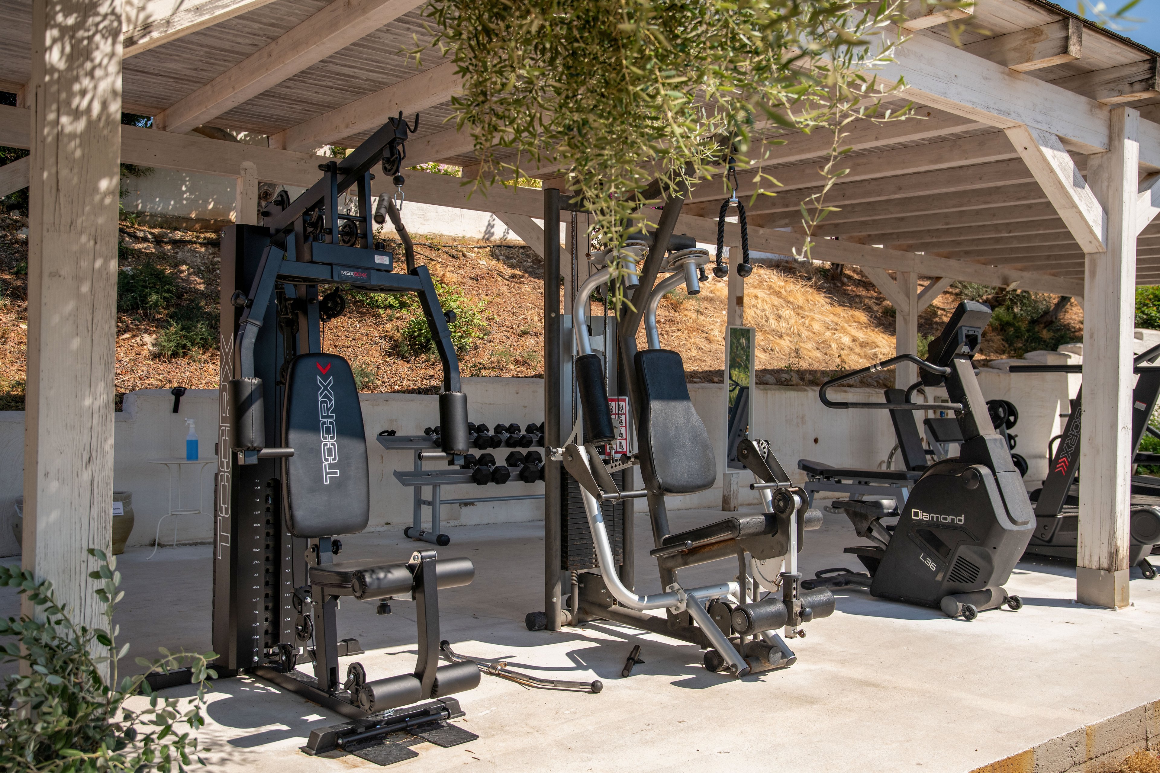 An outdoor gym area features several exercise machines arranged under a wooden pergola. Strength training equipment, including a multi-station weight machine and free weights, stands alongside cardio machines such as an exercise bike. Sunlit trees and a sloped, earthy backdrop frame the open-air workout space. The scene suggests a relaxed yet functional environment for fitness and training.