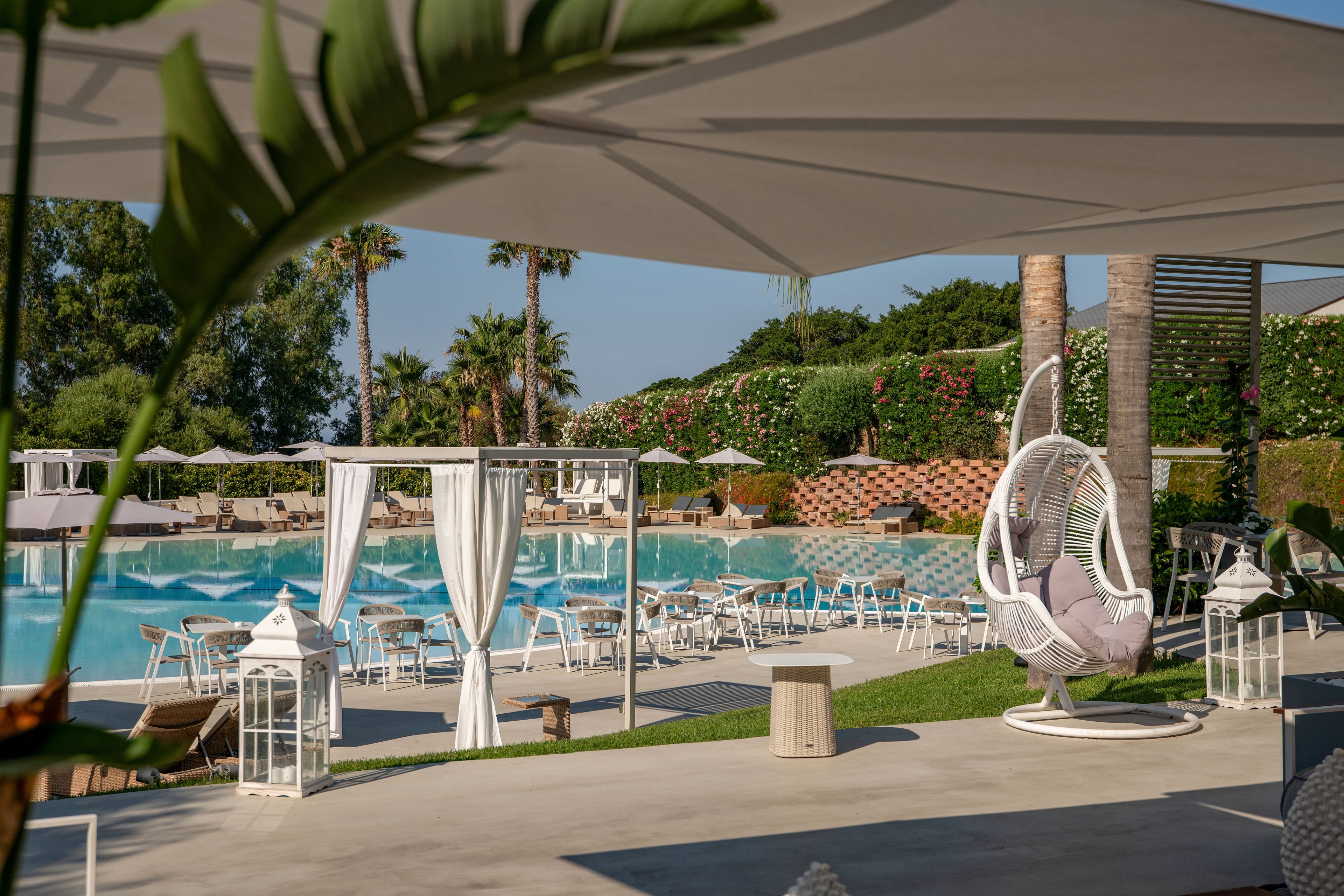 Outdoor resort pool area featuring white cabanas and sun loungers under large shade canopies. A modern hanging chair with cushions sits in the foreground beside decorative lanterns and greenery. The turquoise swimming pool is surrounded by neatly arranged seating and umbrellas, with palm trees and flowering hedges in the background. The scene suggests a relaxed, upscale vacation atmosphere on a sunny day.