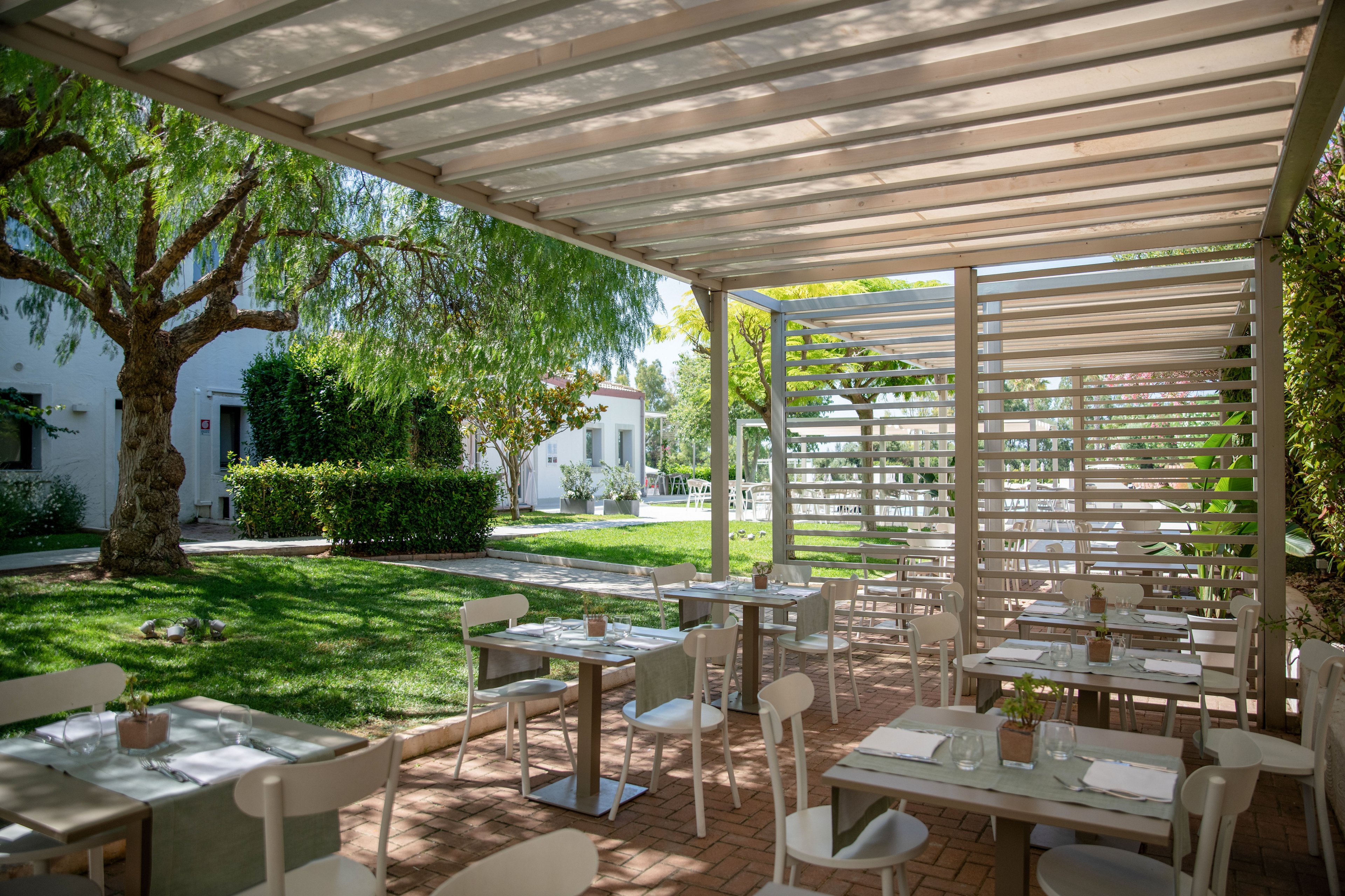 An open-air restaurant terrace features neatly arranged tables and chairs under a modern pergola. The setting overlooks a lush green garden with trees, hedges, and white buildings in the background. Sunlight filters through the slatted roof, creating soft shadows on the brick floor and tabletops. Small plants and tableware add a relaxed, contemporary ambiance to the quiet dining area.