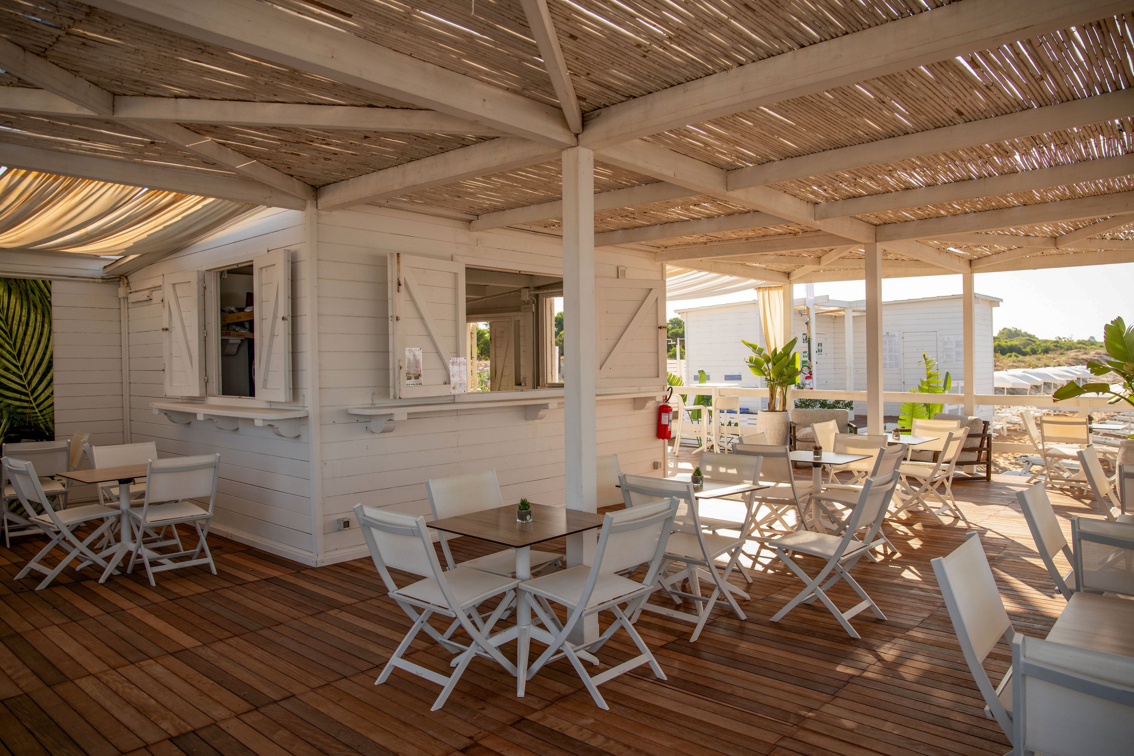 Open-air seaside café with white wooden furniture sits under a rustic pergola roof. Sunlight filters through the slatted canopy onto the wooden deck and neatly arranged tables and chairs. A small white hut with open shutters serves as the bar or service counter, surrounded by potted green plants. The relaxed coastal setting suggests a quiet, inviting spot for outdoor dining and drinks near the beach.