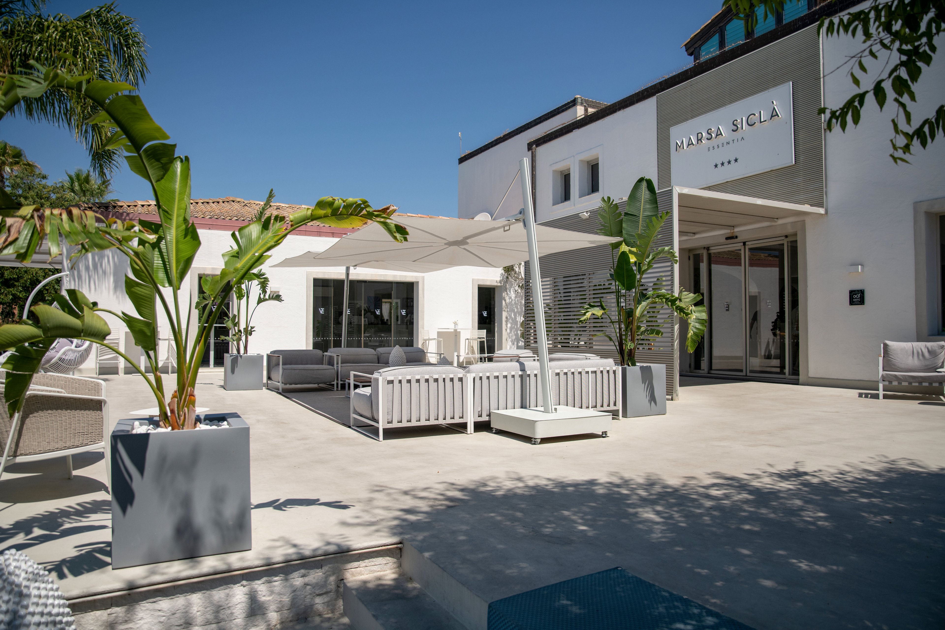 A contemporary white hotel building features an open-air courtyard with stylish lounge furniture and large umbrellas. Palm trees and potted plants add a tropical touch to the clean, minimalist architecture. The entrance area is visible with glass doors and a sign reading "NAPASA SICLA" above. Bright sunlight and clear blue sky create a relaxed, vacation atmosphere.