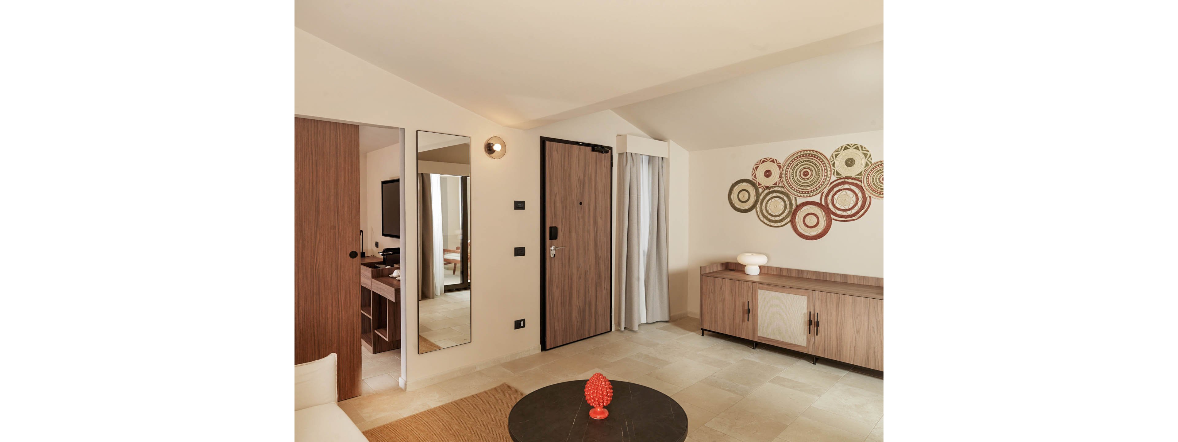 A contemporary hotel room features a wooden entrance door and matching furniture against light beige walls. A sideboard with decorative objects stands beneath circular patterned wall art. A round coffee table sits in the foreground, and a tall mirror reflects a television and desk area. Soft, neutral tones and clean lines create a calm, minimalist atmosphere.