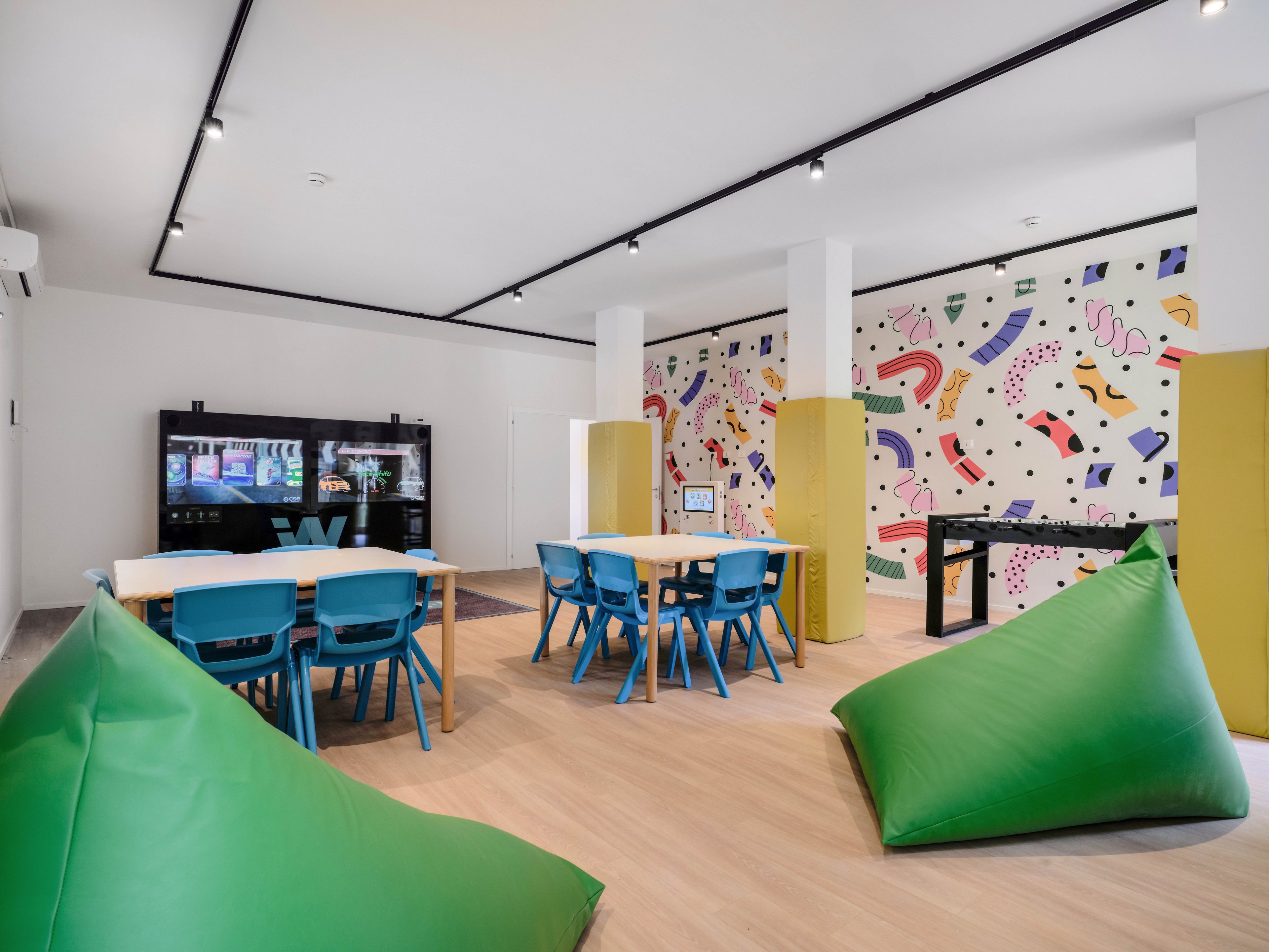 Bright contemporary playroom featuring blue chairs around wooden tables and large green bean bags in the foreground. A wall-mounted screen and digital display suggest an interactive learning or gaming area. The back wall is decorated with a colorful abstract mural, adding a playful atmosphere. The room has light wood flooring, white walls, and ceiling track lighting, creating a clean and inviting space for children or youth activities.