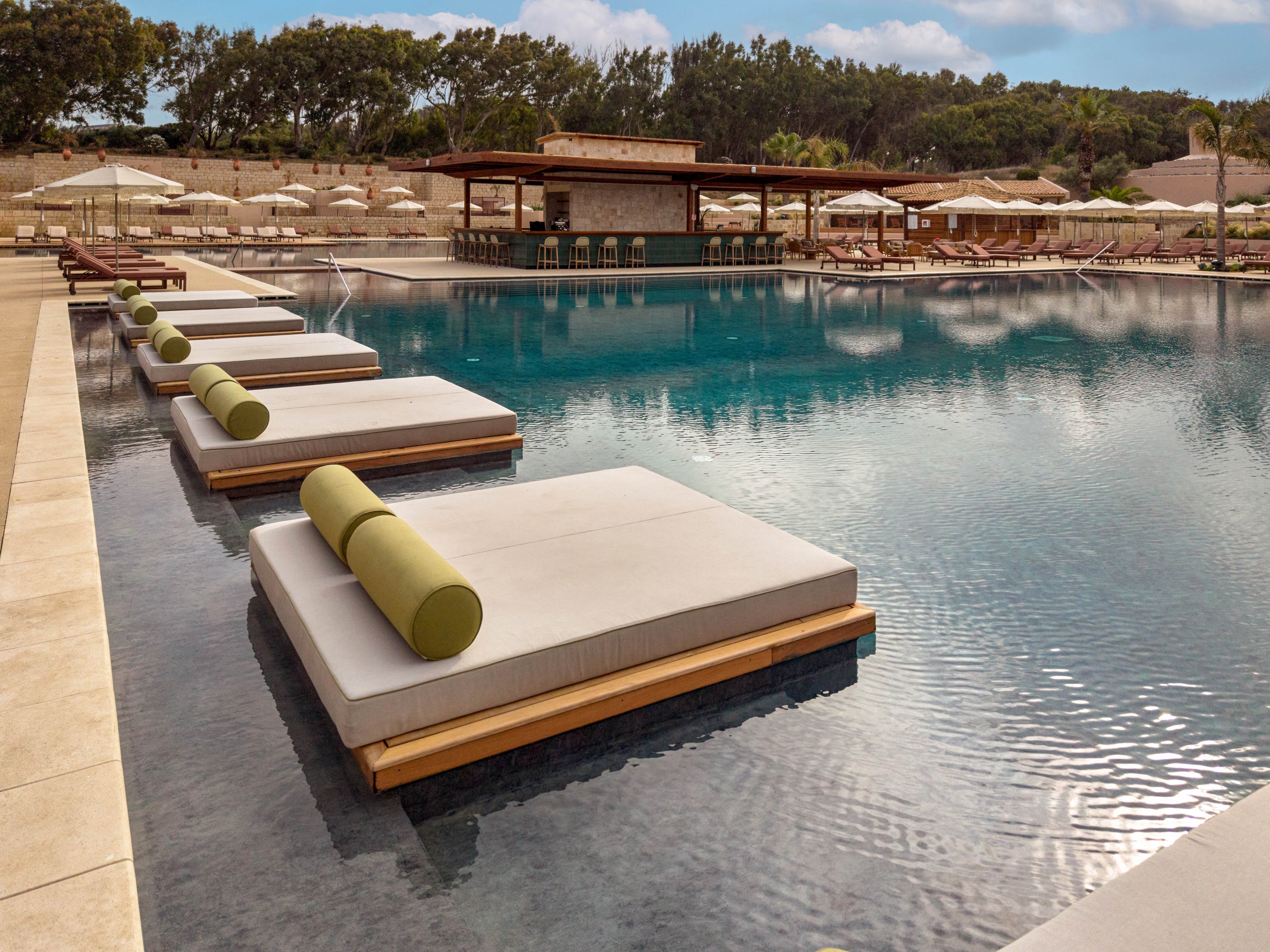 A tranquil resort swimming pool features large cushioned loungers with green bolster pillows set on shallow water. In the background, a wooden pool bar and rows of sun loungers with umbrellas line the deck. Trees and landscaped hills frame the scene under a partly cloudy sky. The overall atmosphere is calm, upscale, and designed for relaxation.