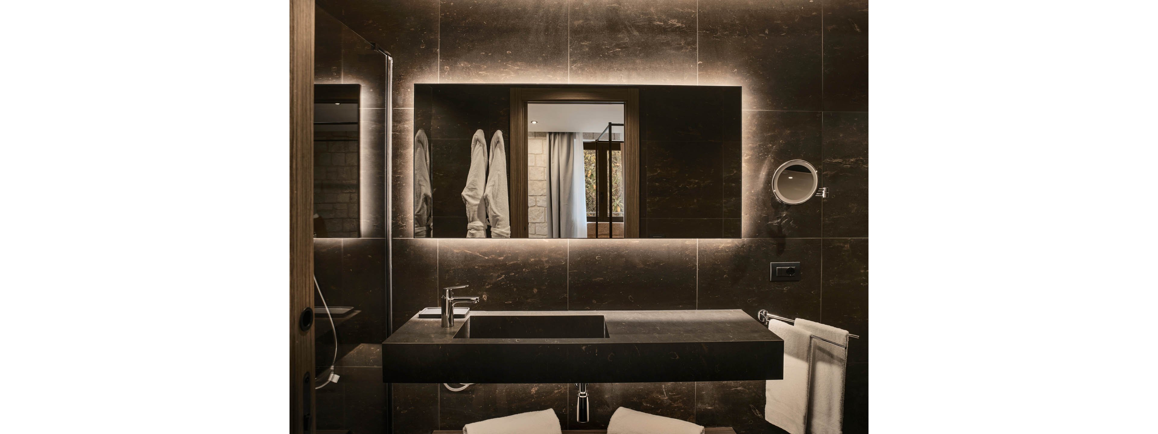 A contemporary bathroom features a large rectangular sink and a wide backlit mirror set against dark stone tiles. Soft lighting highlights the clean lines of the vanity and the wall surfaces. White towels and bathrobes add contrast and a touch of comfort to the otherwise minimalist space. Through the doorway, a glimpse of a curtain and exterior wall suggests a connected bedroom or suite area.