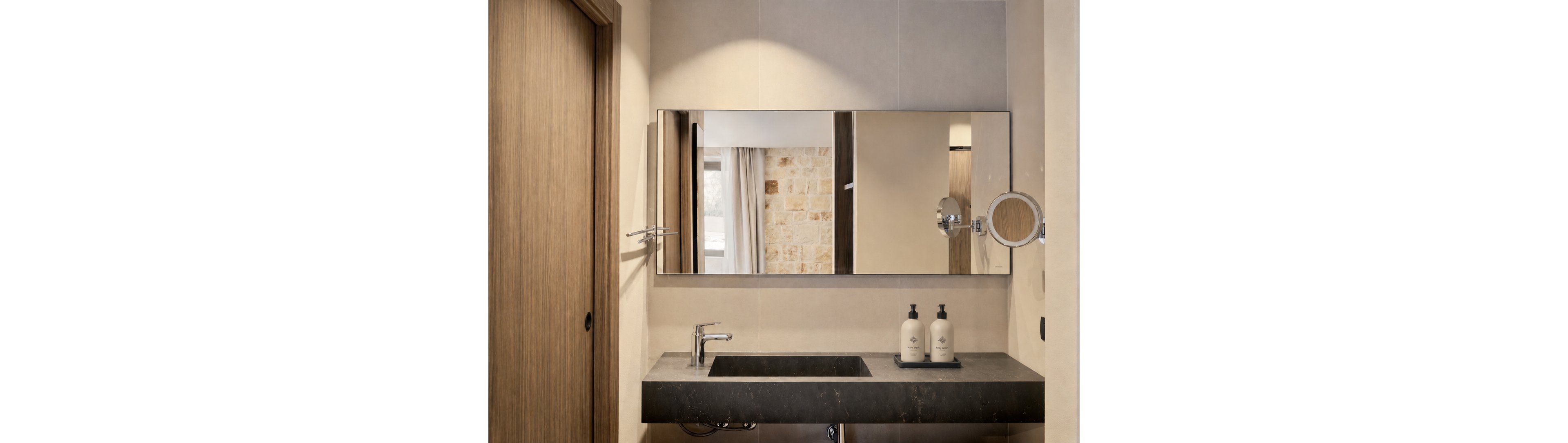 A contemporary bathroom features a wide rectangular mirror above a dark stone vanity. A sleek chrome faucet and integrated sink sit beneath the mirror, with two matching pump bottles arranged on a tray. Warm neutral wall tiles and a wooden sliding door create a minimalist, cozy atmosphere. The mirror reflects a stone accent wall and soft curtains from the adjoining room, adding depth to the space.
