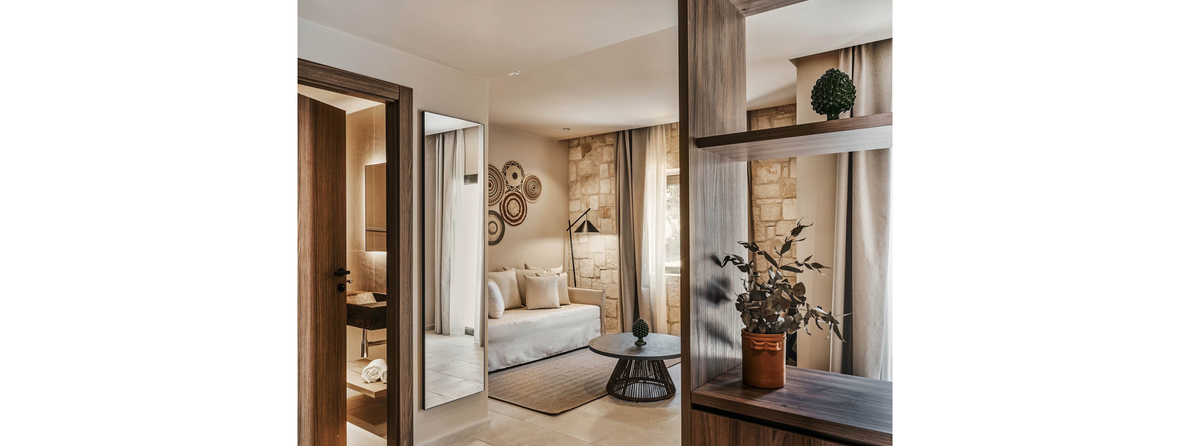A contemporary hotel-style suite features a light-colored sofa bed and soft neutral textiles. Stone accent walls and floor-to-ceiling curtains frame the seating area, while a black floor lamp adds a modern touch. A round coffee table and decorative plants sit in the center, complemented by wooden shelving with potted greenery. Circular wall art and a large mirror near the bathroom entrance complete the stylish, inviting interior.