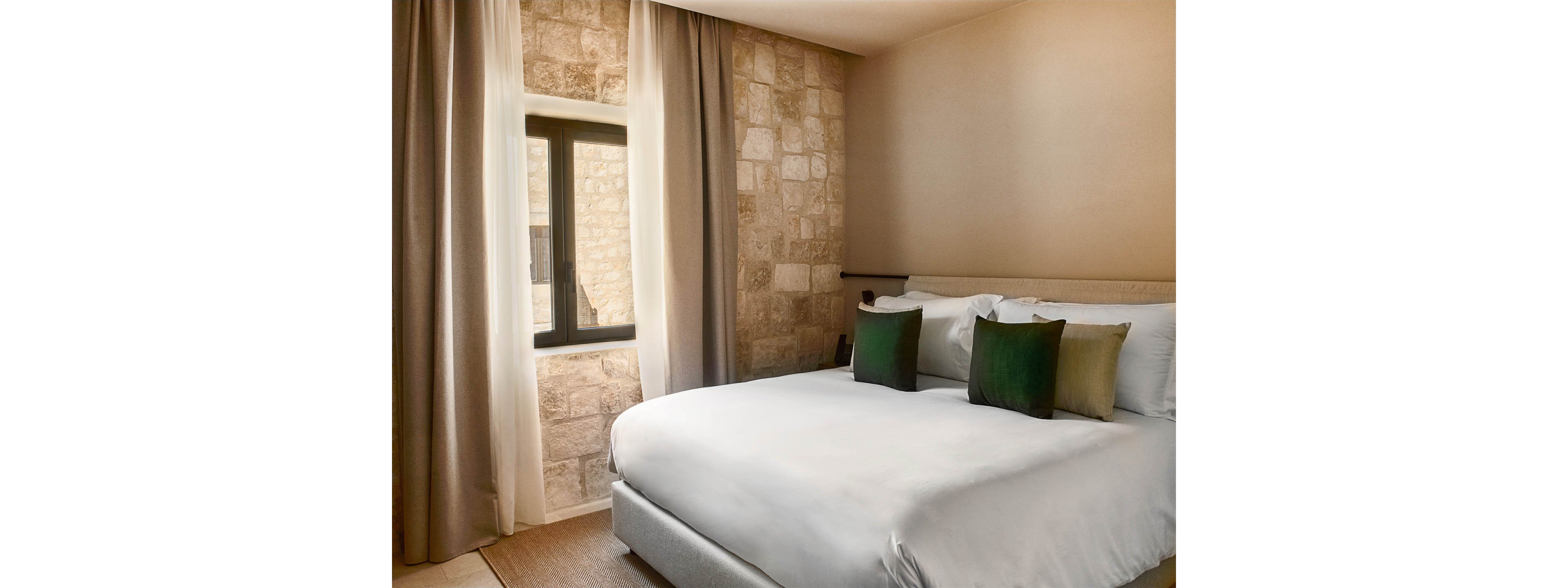 A neatly made bed with white linens and green accent pillows sits in a modern bedroom. One wall features a textured stone finish, contrasting with smooth beige walls. Natural light enters through a small rectangular window framed by long neutral curtains. The overall scene feels calm, minimal, and inviting, suitable for a hotel or contemporary home interior.