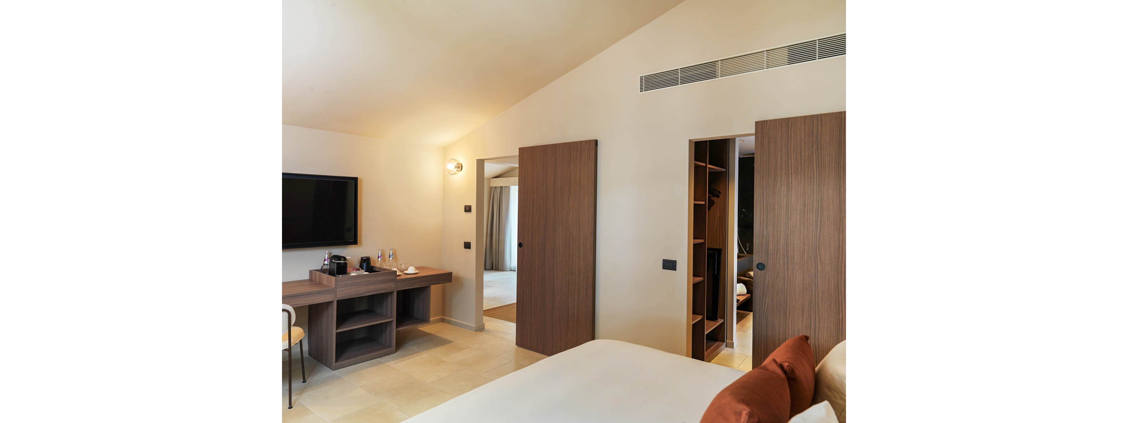 A contemporary hotel room features a neatly made bed with rust-colored accent pillows in the foreground. Along the wall, a flat-screen television hangs above a wooden desk holding a coffee station and glassware. Two wooden sliding doors reveal a walk-in wardrobe and an adjacent room with curtains. Neutral walls, tiled flooring, and soft lighting create a clean, minimalist atmosphere.