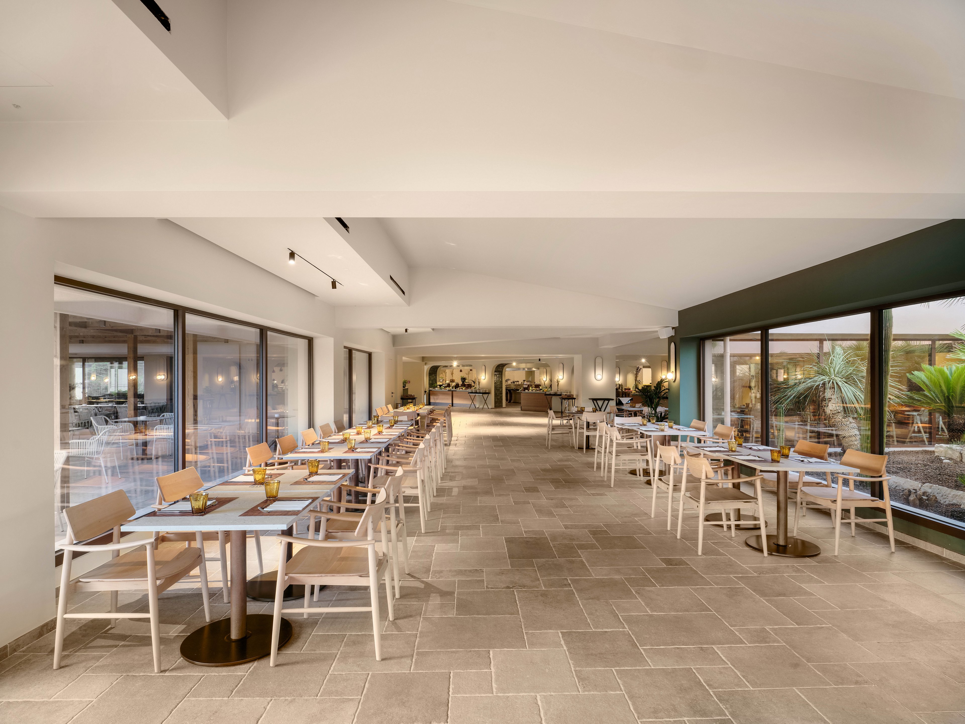 Spacious contemporary restaurant dining area with light wooden tables and chairs arranged in neat rows. Large windows on one side provide natural light and views of an outdoor terrace with plants. Neutral tones, clean lines, and minimalist decor create an airy, relaxed atmosphere. The far end of the room features a service or buffet area, enhancing the hospitality setting.