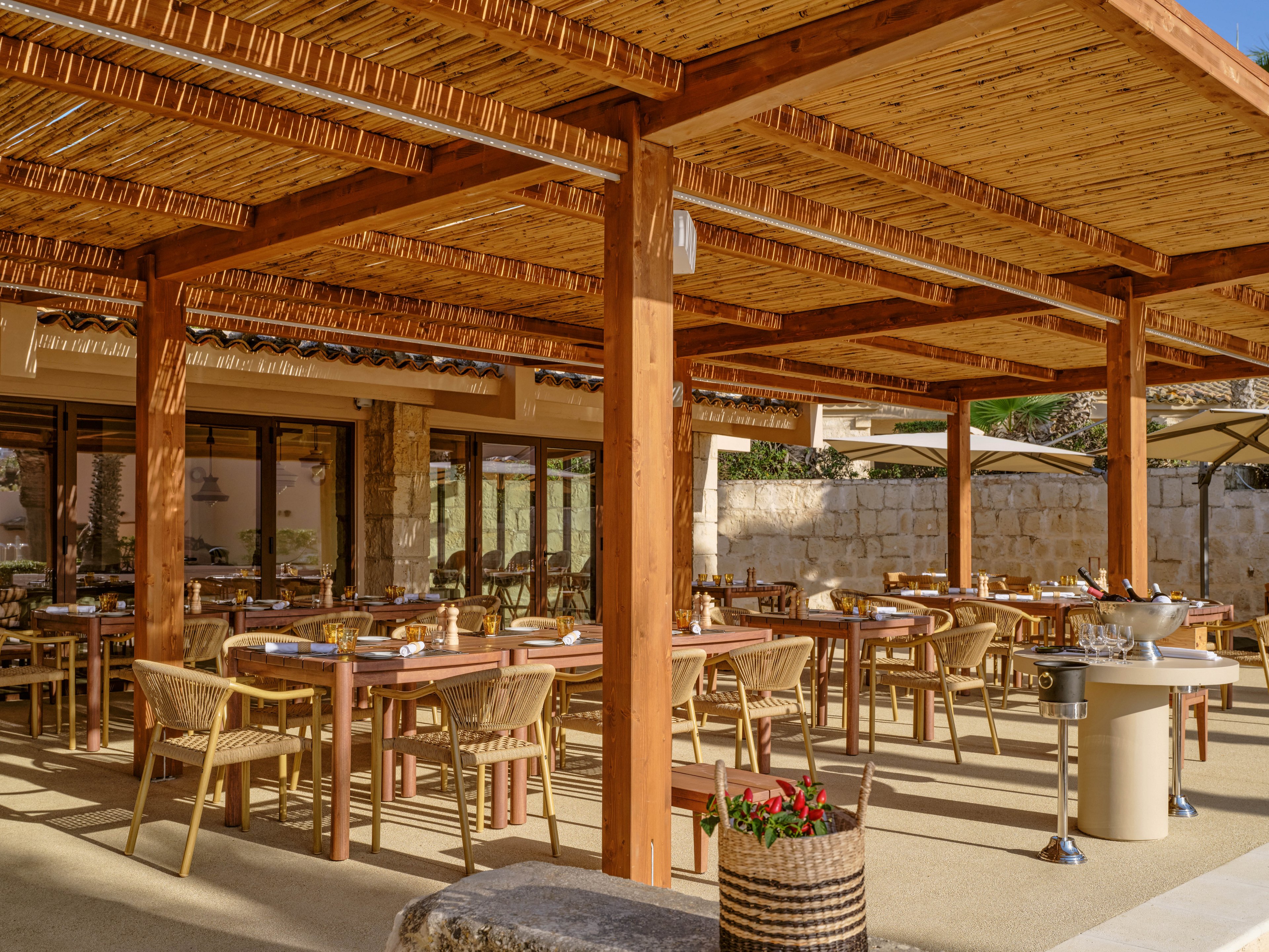 An inviting outdoor restaurant terrace features wooden tables and woven chairs arranged beneath a warm-toned wooden pergola. Sunlight fills the space, highlighting neatly set tables with plates, glasses, and condiments ready for guests. Stone walls and large glass doors frame the background, suggesting a Mediterranean or resort-style setting. The overall atmosphere is relaxed and welcoming, ideal for al fresco dining.