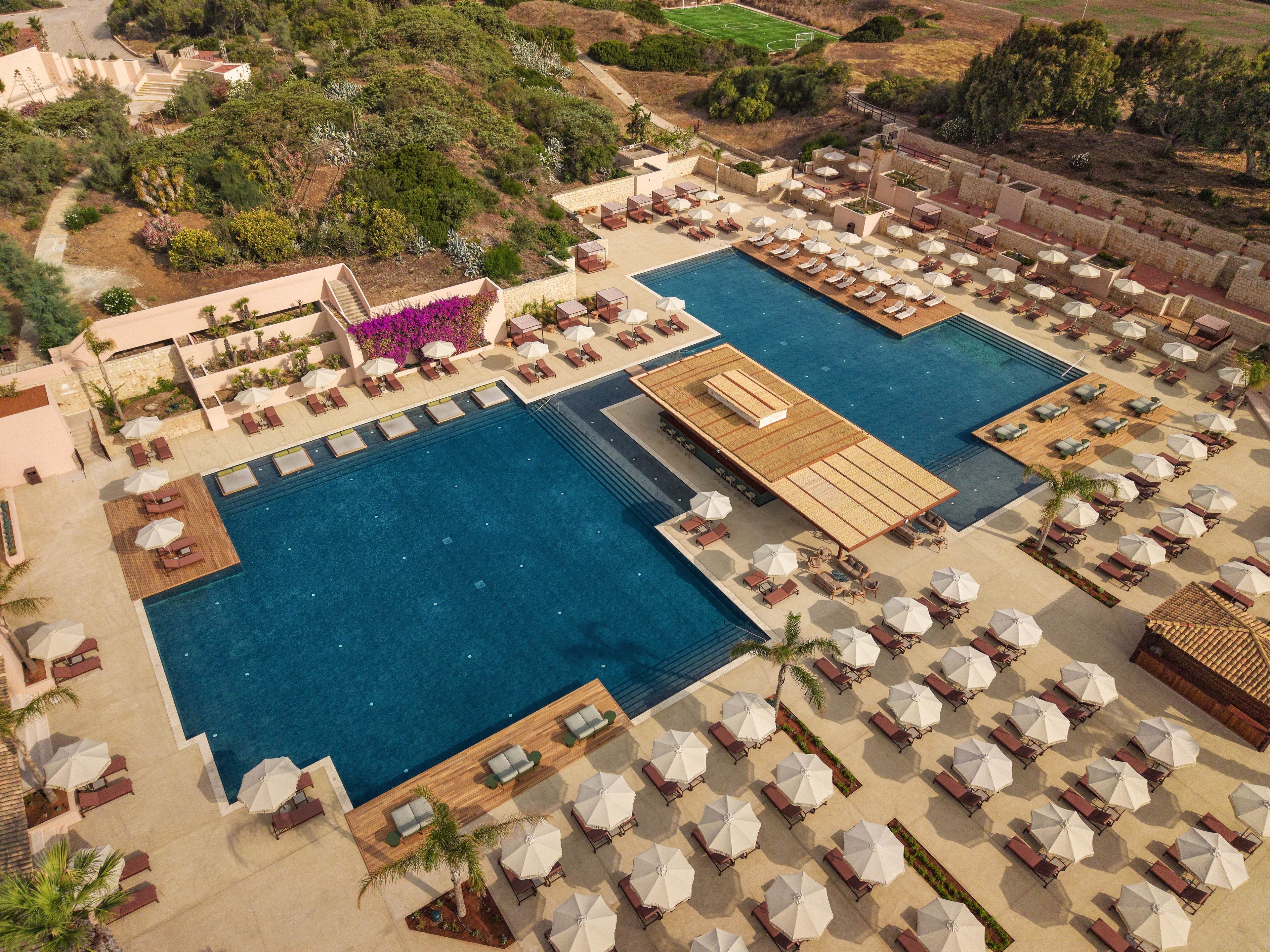 A large resort swimming pool complex is seen from above, featuring multiple rectangular pools surrounded by sun loungers and umbrellas. The central pool area includes a wooden deck structure extending over the water, creating a focal point for relaxation. Beige stone paving, neatly arranged seating, and landscaped greenery frame the pools, giving the scene an organized and upscale appearance. In the background, gardens and natural vegetation blend the resort into the surrounding landscape.