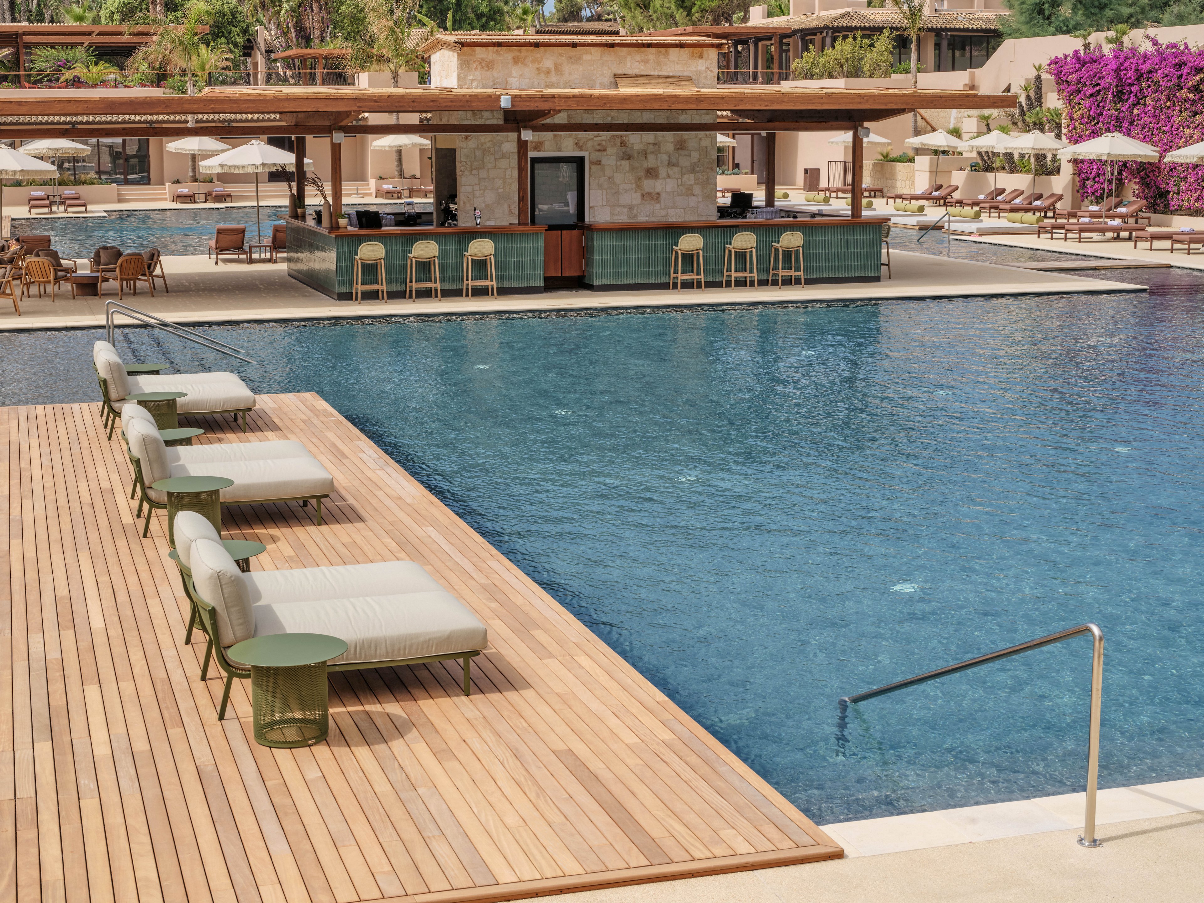 A large resort swimming pool is surrounded by a wooden deck with cushioned sun loungers and small side tables. In the background, an open-air pool bar with stools sits at the center of the water, shaded by a flat roof. Umbrellas and outdoor seating areas line the far side of the pool, framed by lush greenery and flowering vines. The scene suggests a relaxing vacation atmosphere in a warm, sunny climate.