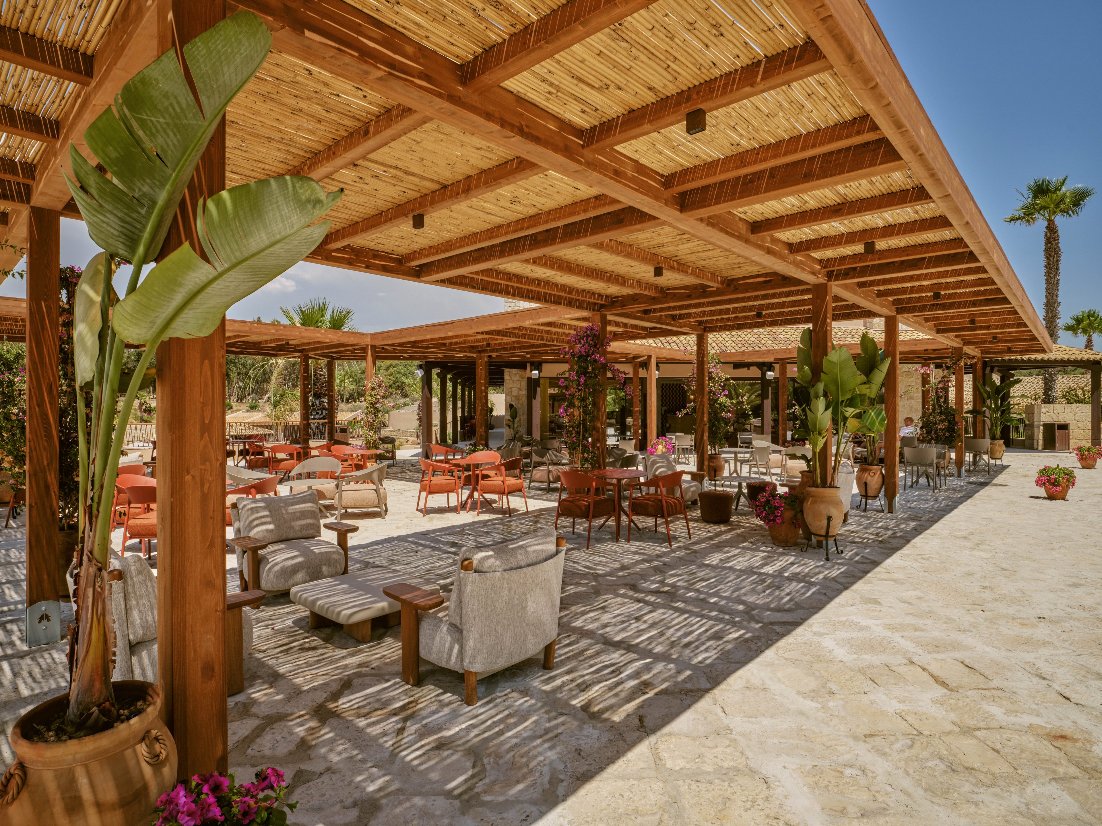 Open-air terrace with wooden pergolas shelters a variety of lounge chairs and dining tables. The setting appears to be a warm Mediterranean or resort-style courtyard with stone flooring. Potted tropical plants and flowering vines add color and texture around the seating area. Sunlight filters through the slatted roof, casting striped shadows across the ground and furniture.