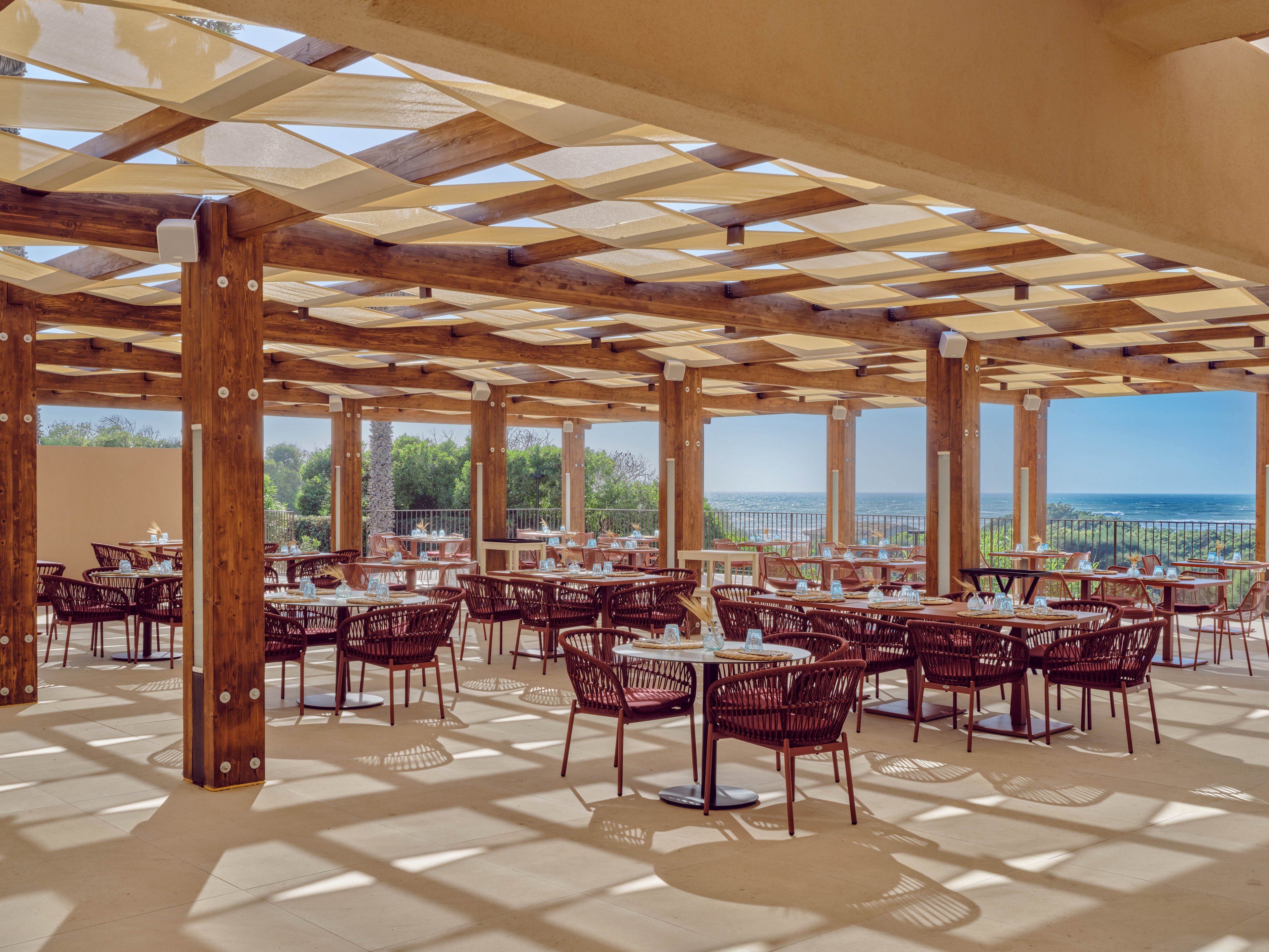 An open-air restaurant features rows of tables and wicker-style chairs beneath a wooden pergola casting geometric shadows on the floor. The setting overlooks a scenic coastline with blue water and greenery visible beyond a railing. Sunlight filters through the patterned roof, creating a warm and inviting atmosphere. The space appears ready for guests, with table settings arranged neatly in a relaxed outdoor dining area.