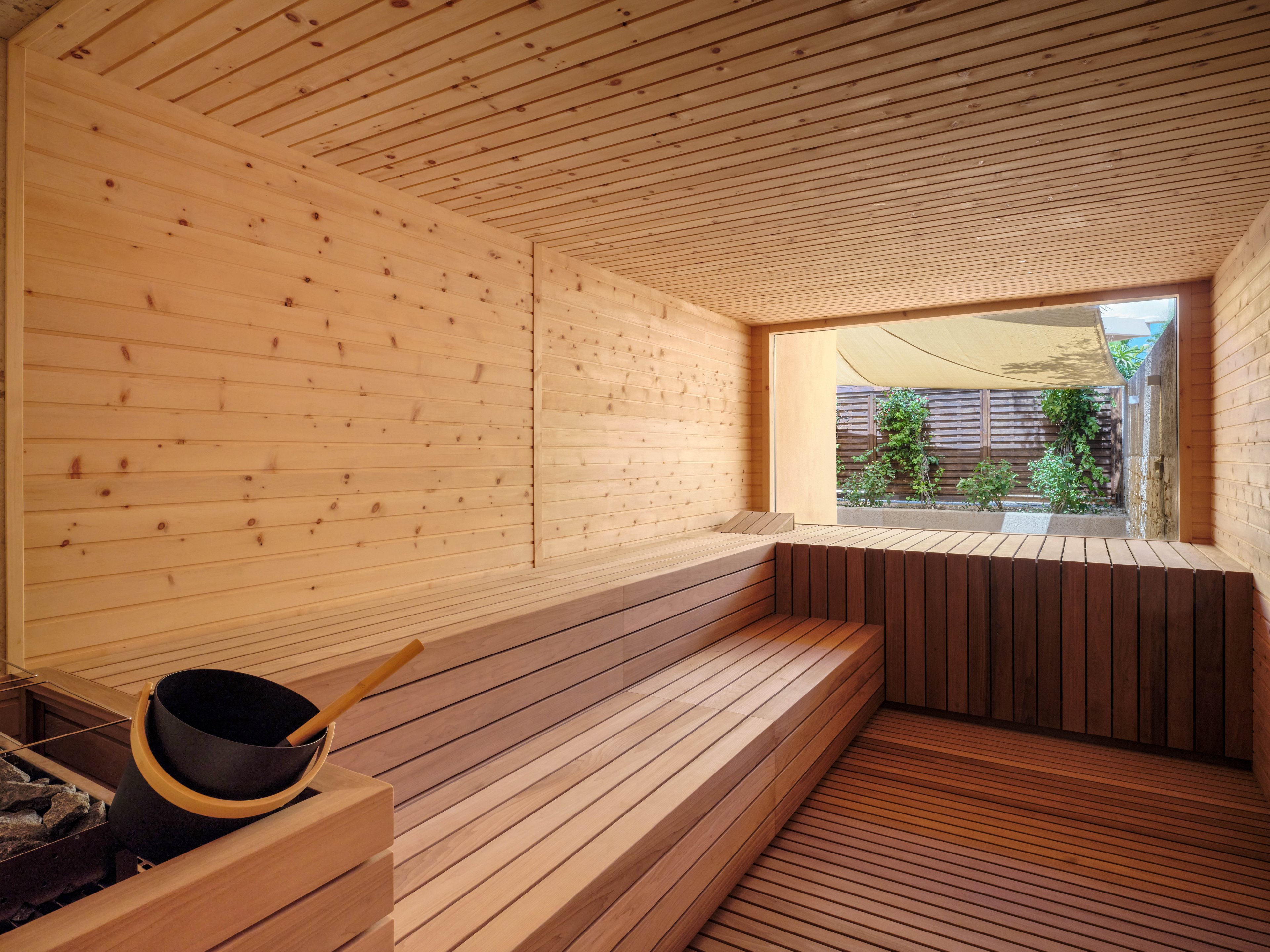 A contemporary wooden sauna interior features tiered bench seating and clean, minimalist lines. Light-toned wood covers the walls, ceiling, and floor, creating a warm and natural atmosphere. A sauna bucket and ladle sit near the heater area, suggesting a relaxing spa experience. Large window openings reveal an outdoor garden with greenery and wooden fencing, adding a tranquil, nature-inspired backdrop.