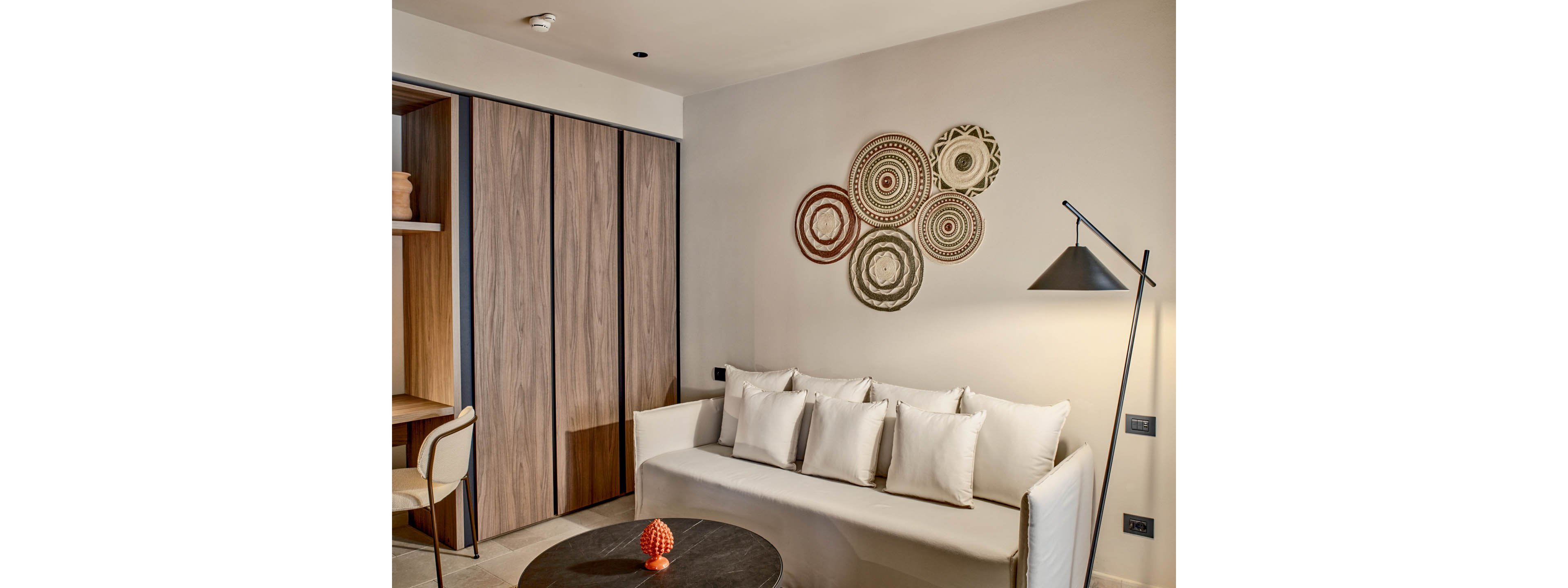 Contemporary living room interior featuring a light-colored sofa with multiple white cushions. Circular decorative wall art hangs above the sofa, adding texture and visual interest. A black floor lamp stands beside the seating area, while a wooden wardrobe and small desk are visible to the side. The overall design is minimalist with warm neutral tones and subtle accent decor.