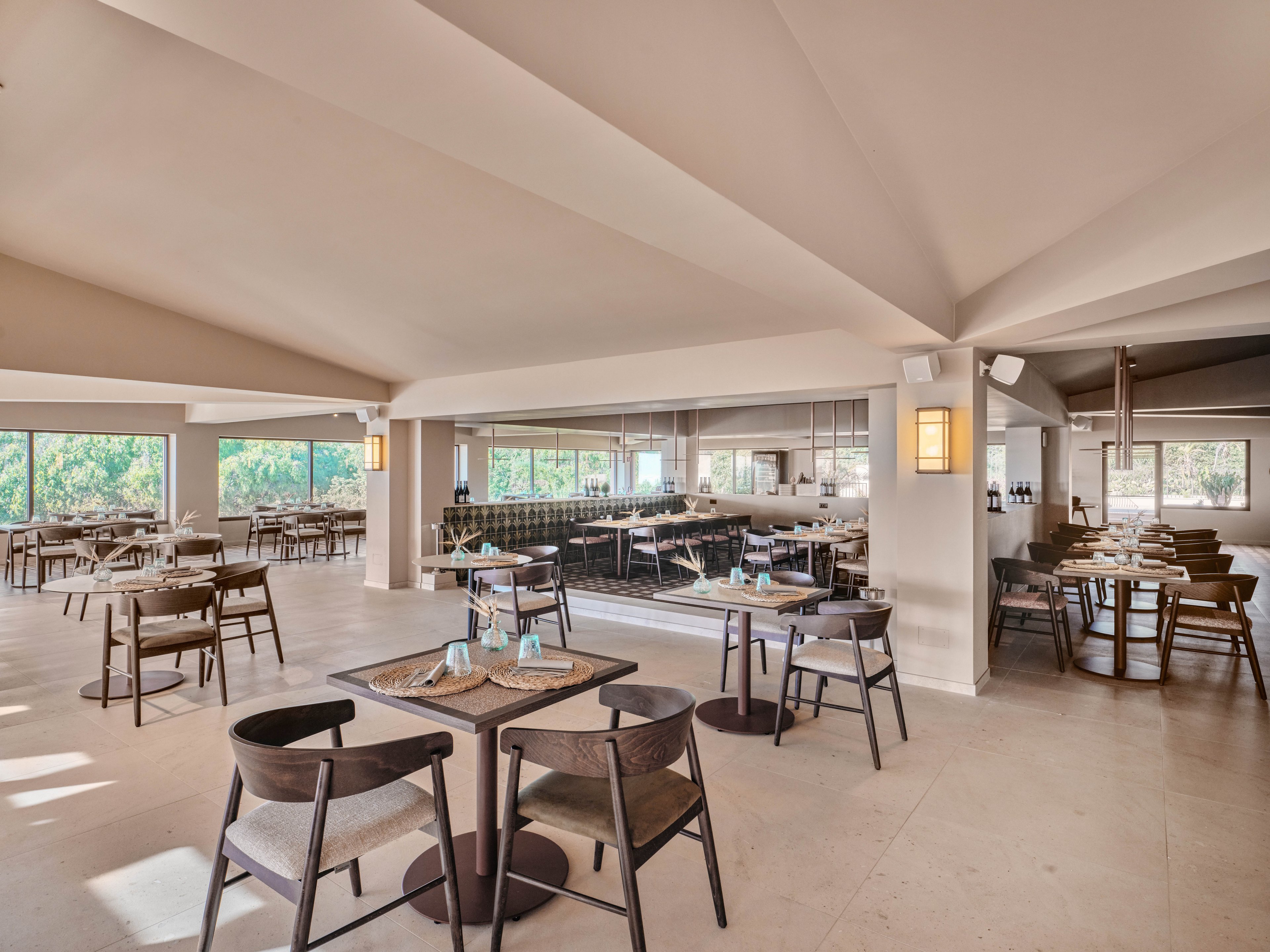 Spacious contemporary restaurant dining area with neatly arranged wooden tables and chairs. Large windows and glass walls let in natural light and reveal greenery outside. Neutral beige tones, clean lines, and minimalist decor create a calm, airy atmosphere. Table settings include placemats, napkins, and small decorative items, suggesting readiness for guests.