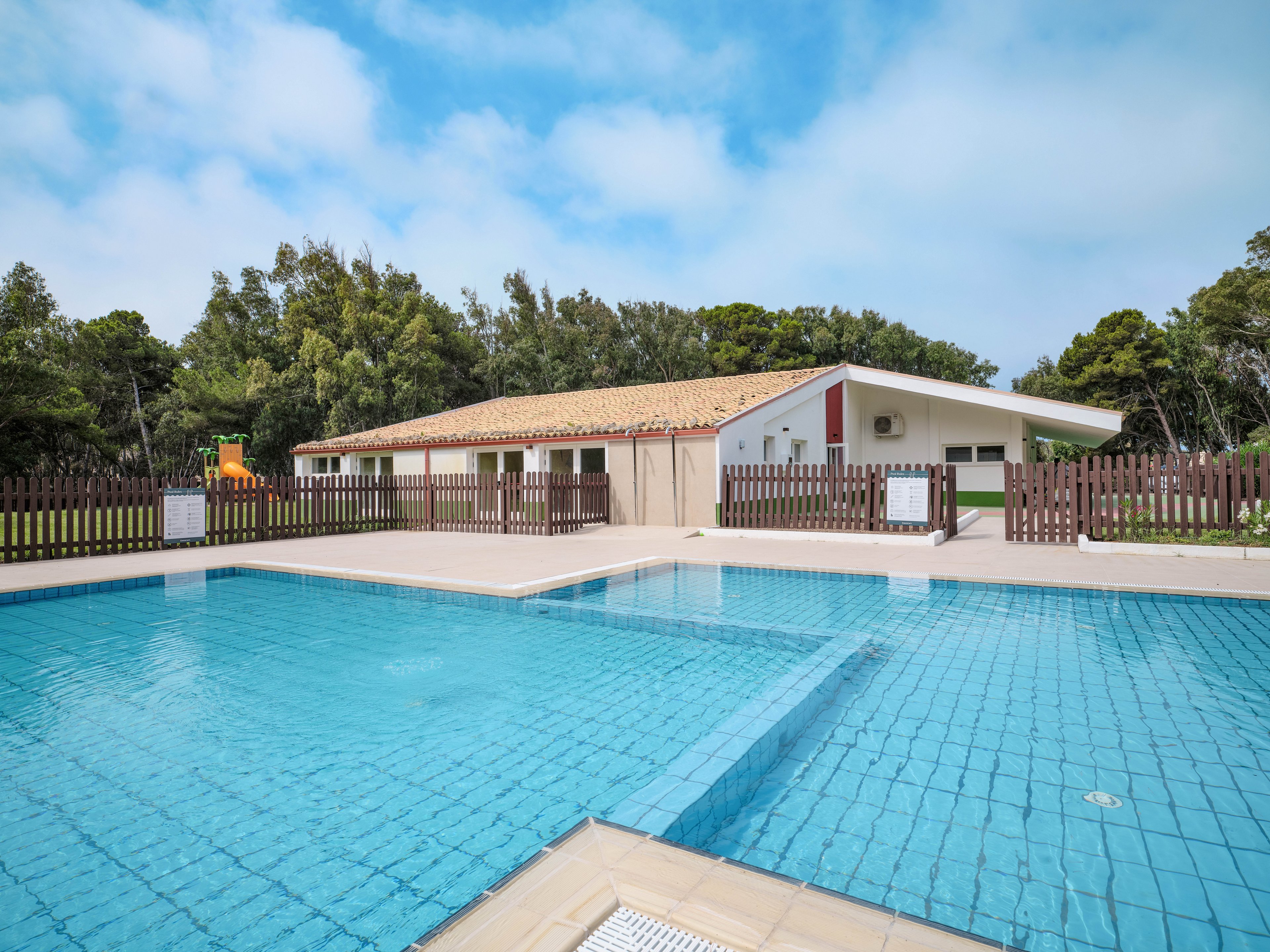 An outdoor swimming pool with clear blue water is shown in the foreground, surrounded by a light-colored deck. Behind the pool stands a low, modern lodge-style building with a tiled roof and white walls. A wooden fence encloses the area, with trees and greenery visible beyond it. The sky is bright with scattered clouds, creating a calm and inviting vacation atmosphere.