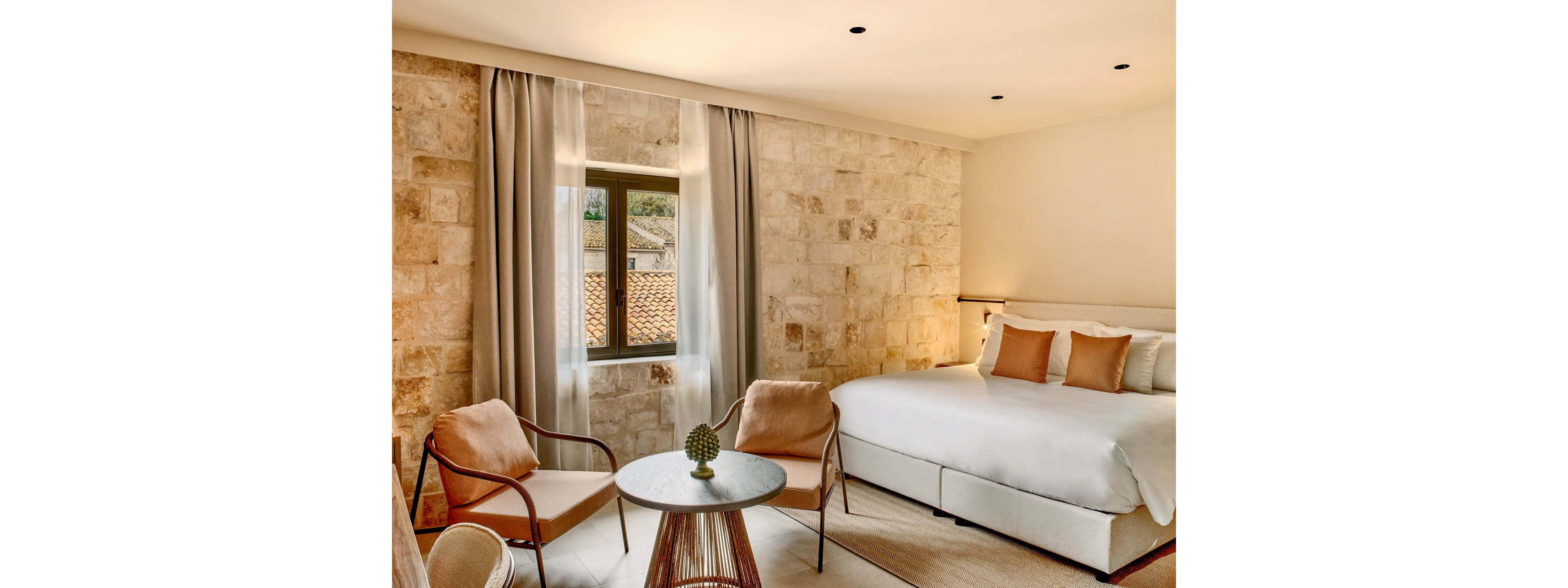 A warm, inviting hotel bedroom features a large white bed with multiple beige and peach cushions. The room has textured stone walls and a window overlooking tiled rooftops, framed by long neutral curtains. A small round table with a decorative object is surrounded by two modern armchairs, creating a comfortable seating area. Soft, natural light and minimal decor contribute to a calm, contemporary atmosphere.