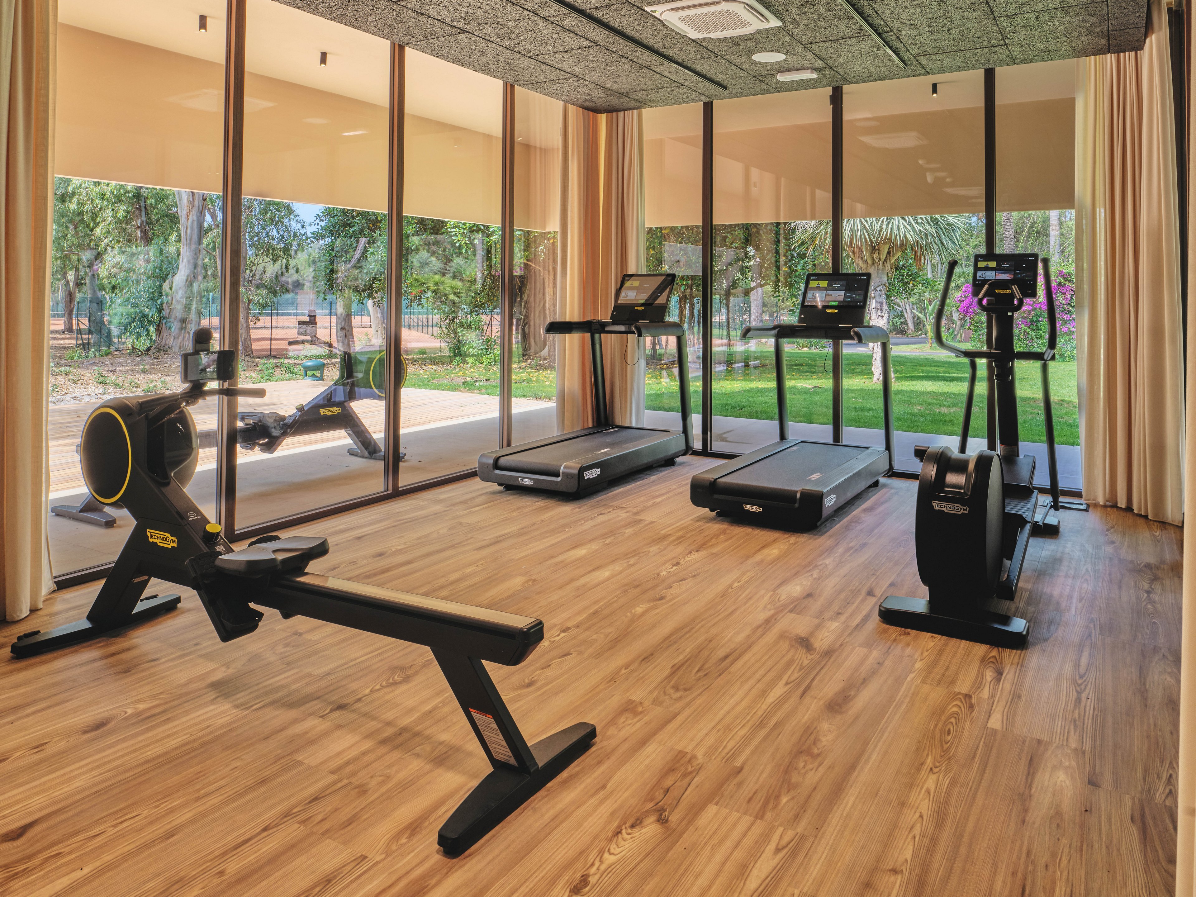 A bright fitness room features a rowing machine, two treadmills, and an exercise bike arranged on a wooden floor. Large floor-to-ceiling windows reveal a lush outdoor garden with trees and greenery. Neutral walls and ceiling panels create a clean, contemporary atmosphere. Natural light fills the space, emphasizing the warm tones of the wood and the sleek black gym machines.