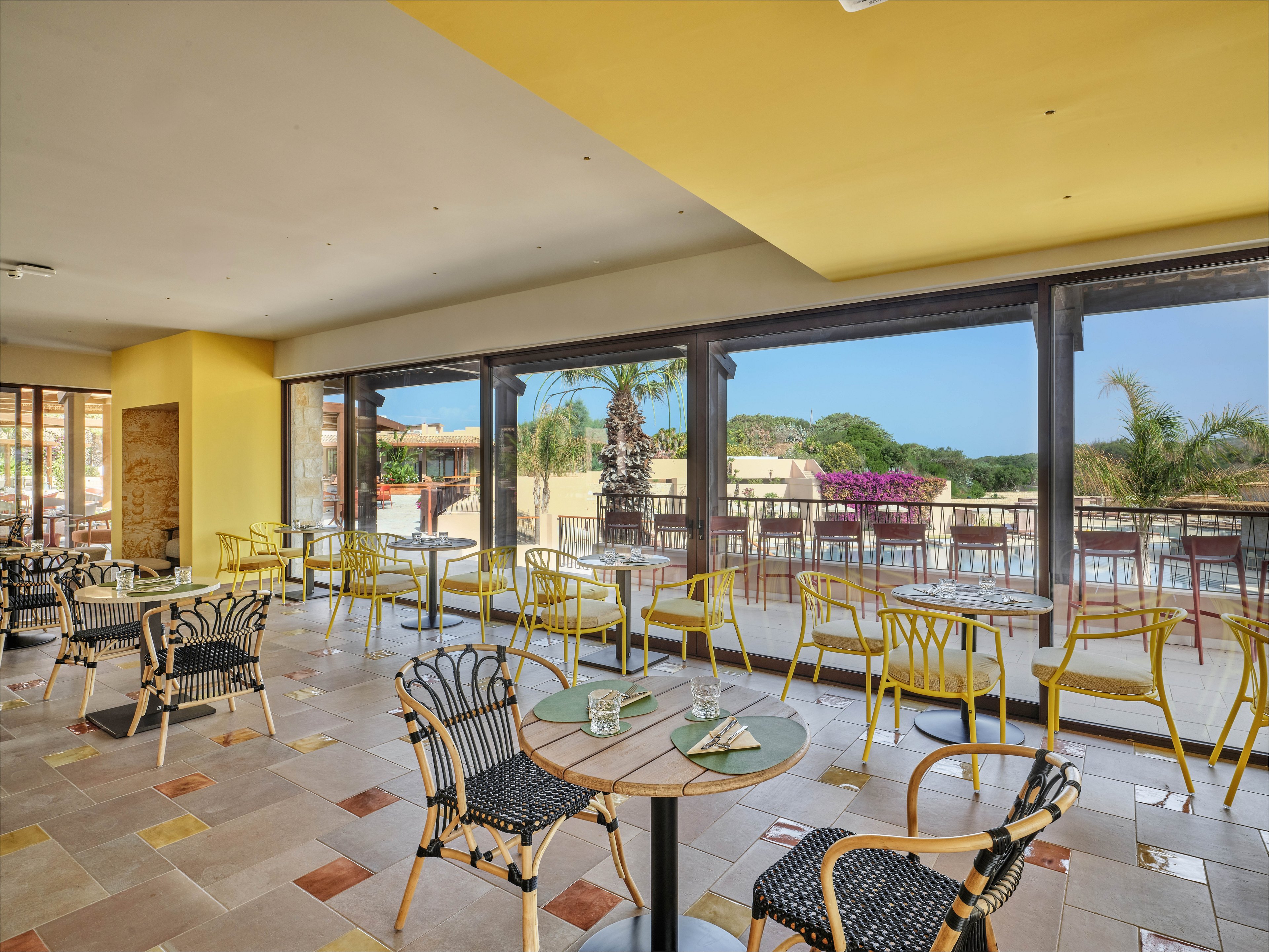Bright indoor café seating overlooks a spacious outdoor terrace at a resort. Yellow walls and large floor-to-ceiling windows create an airy, sunlit atmosphere. Wicker chairs and small round tables are arranged neatly, with glasses and plates set for guests. Outside, palm trees, flowering plants, and distant hills enhance the relaxed vacation setting.