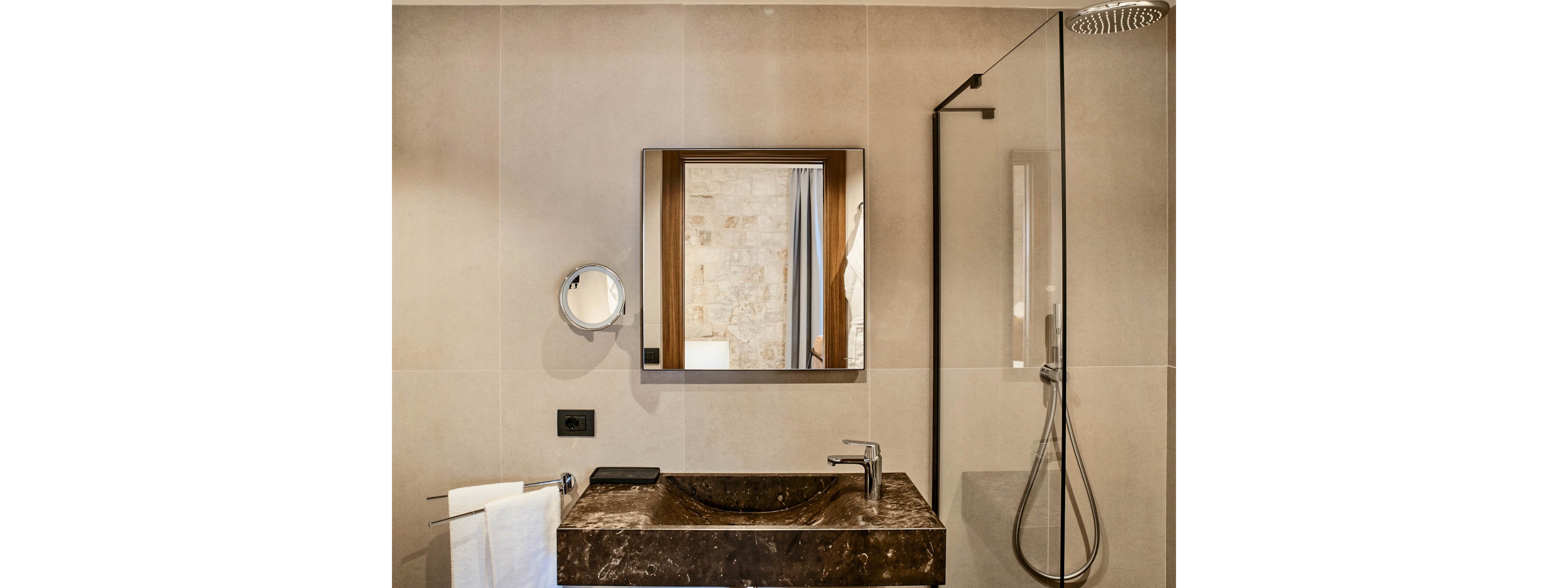 A contemporary bathroom features a dark marble sink vanity with a sleek chrome faucet. Above the vanity hangs a square mirror, reflecting a neutral-toned wall and doorway. To the right, a glass shower enclosure with a handheld showerhead adds to the minimalist design. A round magnifying mirror and neatly folded white towels complete the clean, upscale look.