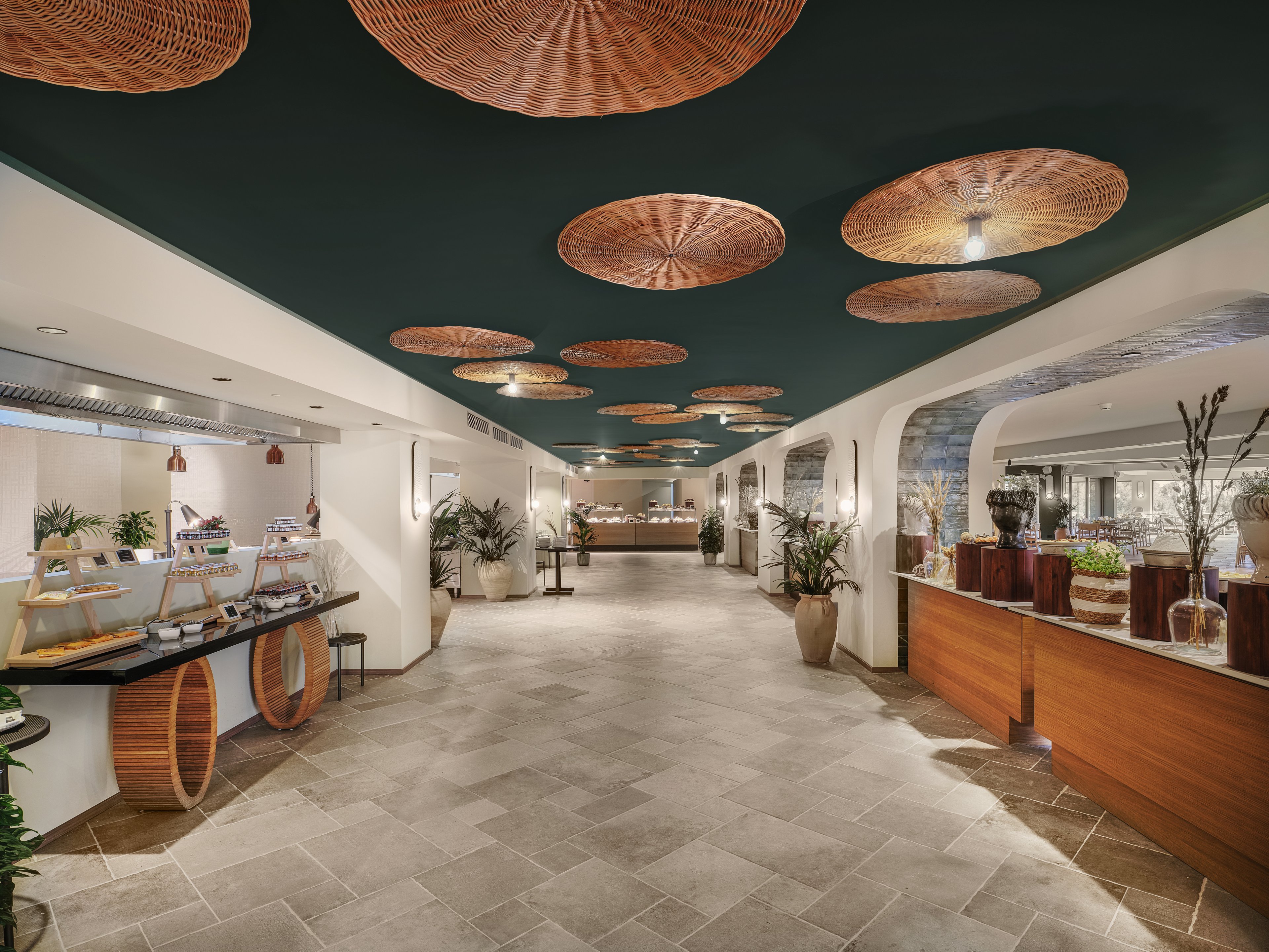 Spacious interior corridor featuring a contemporary buffet area in a hotel or resort. The setting includes food stations with wooden accents and decorative baskets hanging from a dark ceiling. Neutral stone flooring and curved white walls create an elegant, minimalist atmosphere. Potted plants and vases add natural elements to the refined dining environment.