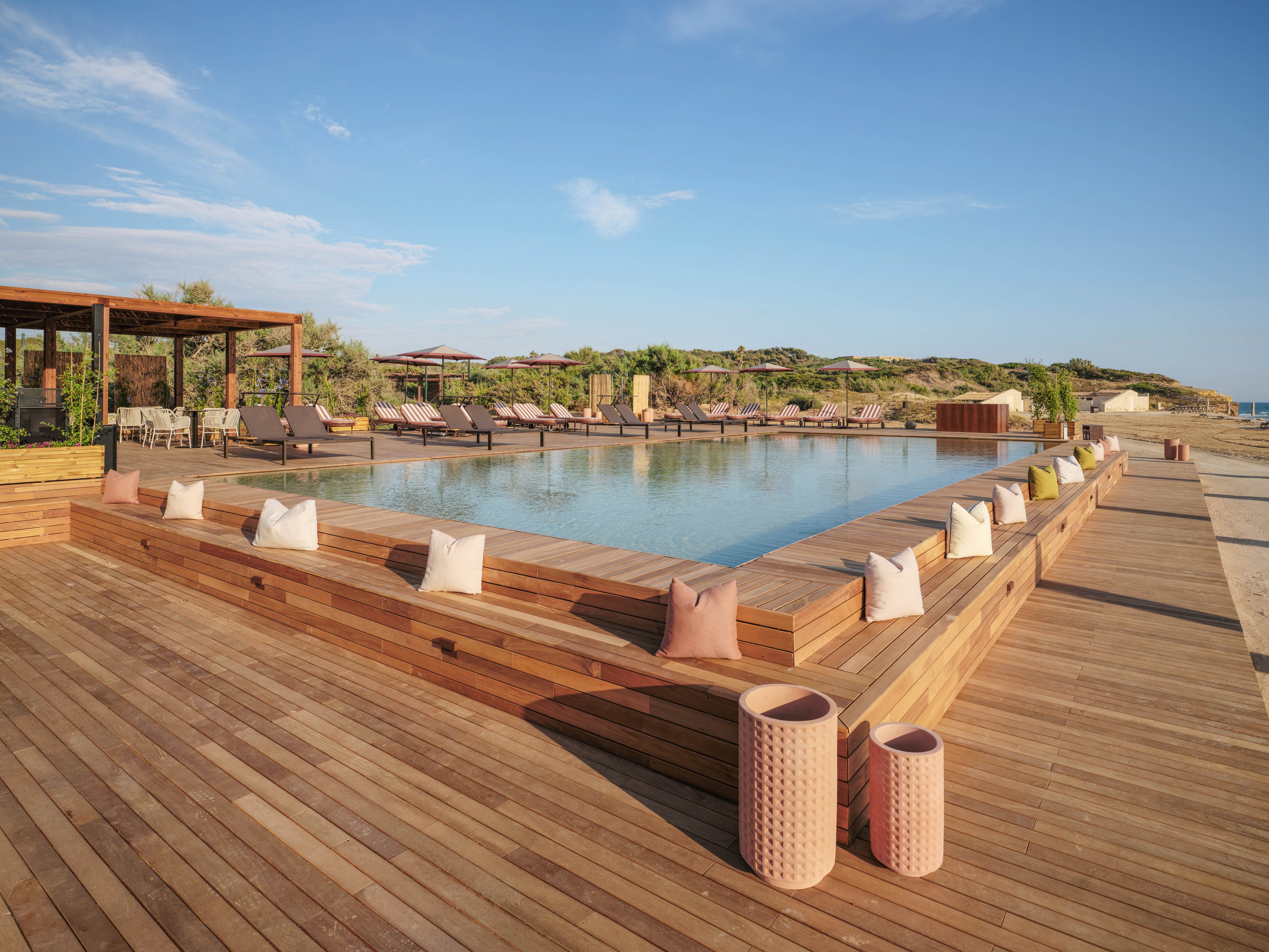 An elegant infinity-style pool is surrounded by a spacious wooden deck at a beachfront resort. Lounge chairs and umbrellas line the far side of the pool, facing toward the ocean and sandy dunes. Cushions are arranged along the deck edges, and decorative cylindrical stools sit in the foreground. The scene is bathed in warm natural light under a clear blue sky, creating a tranquil vacation atmosphere.