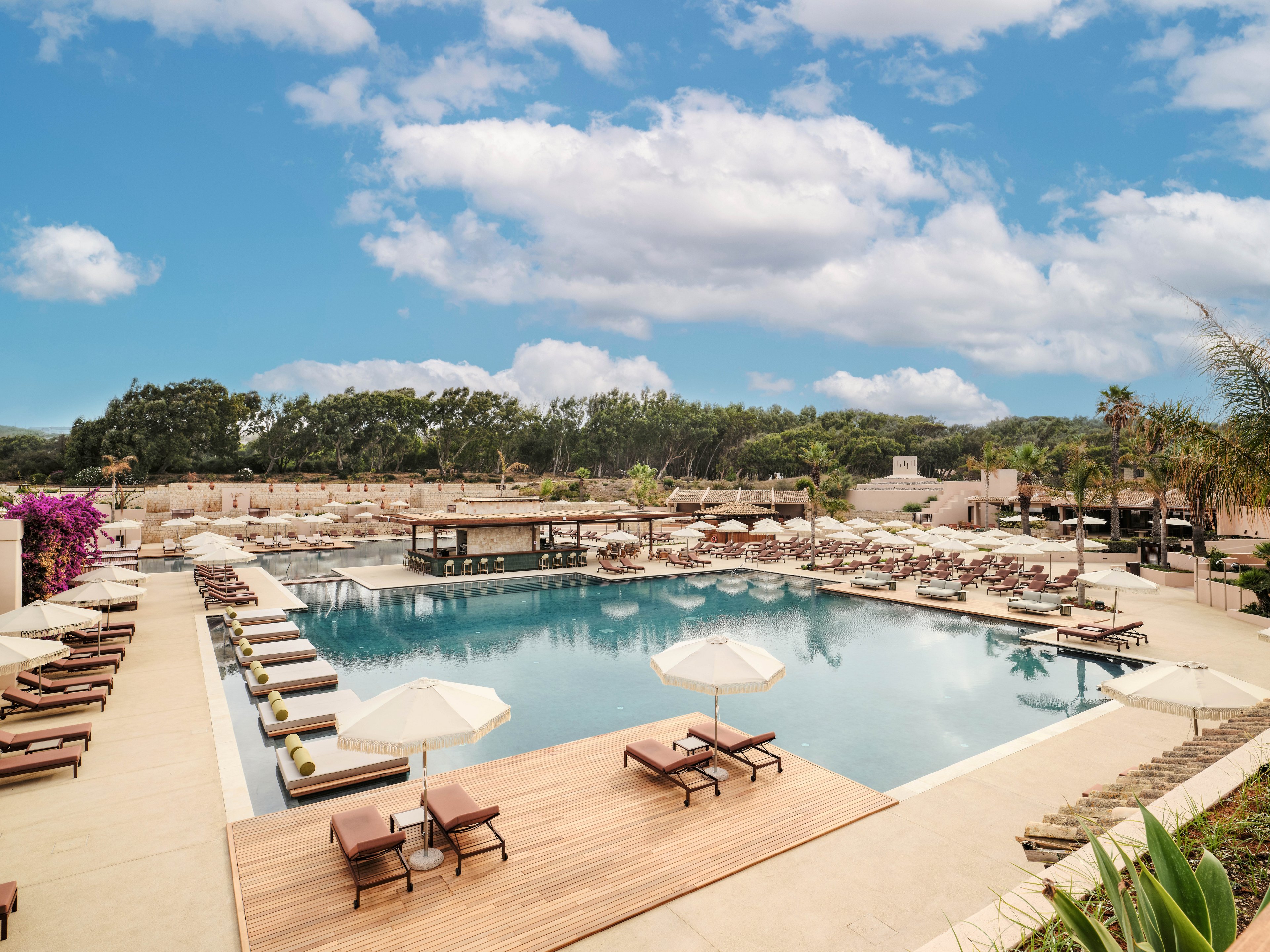 A large outdoor swimming pool is surrounded by numerous sun loungers and umbrellas at a modern resort. Wooden decking and light-colored stone surfaces create a clean, upscale atmosphere. Lush greenery and trees frame the background under a bright blue sky with scattered clouds. The scene suggests a relaxing vacation setting focused on leisure and comfort.