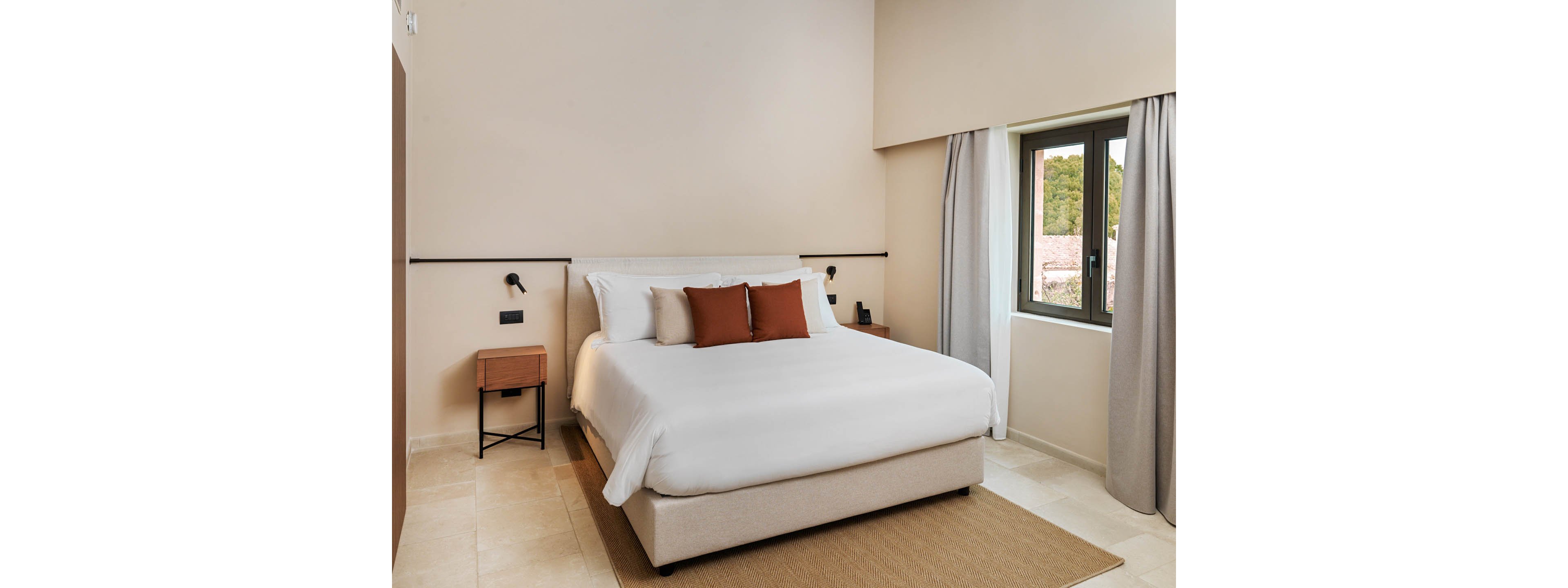 A neatly made double bed with white linens and brown accent pillows stands in a modern minimalist bedroom. The room features light beige walls, a small wooden nightstand, and simple black wall-mounted reading lights. Natural light enters through a large window with gray curtains, revealing greenery and rooftops outside. A neutral rug and tiled floor complete the clean, contemporary look.
