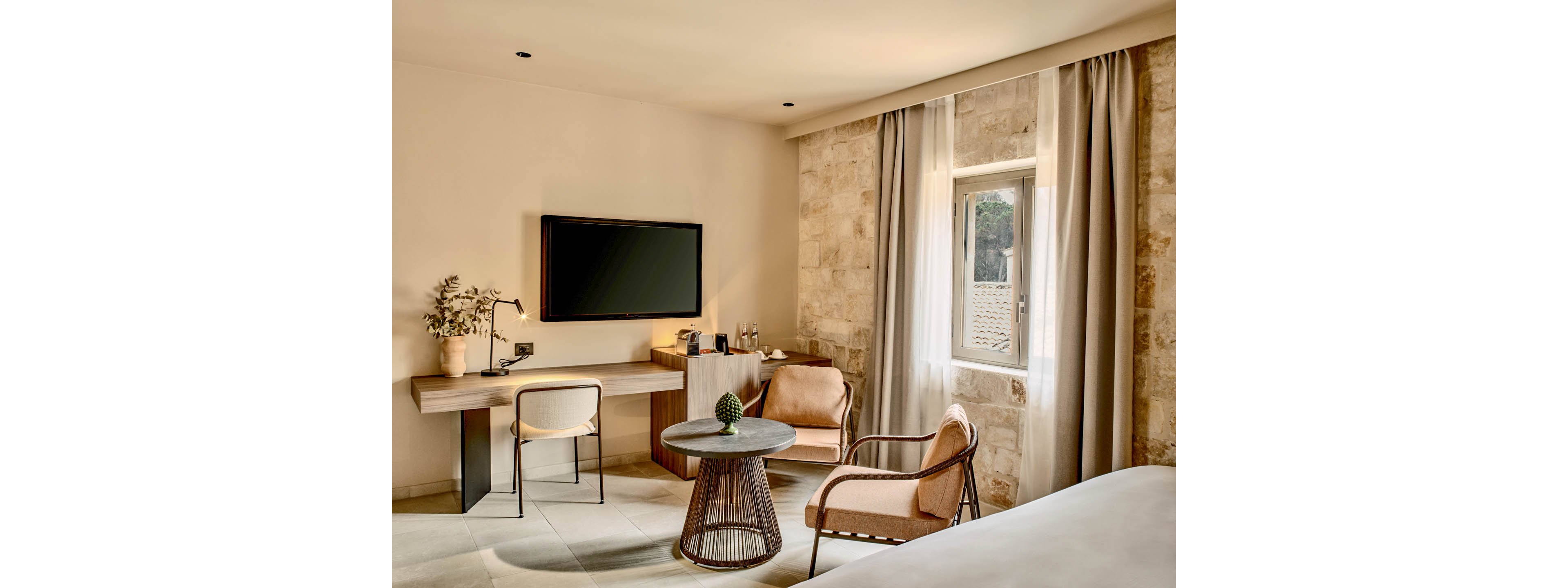 A contemporary hotel room features a wall-mounted flat-screen television above a minimalist desk with a chair. A small round table and an upholstered armchair create a cozy seating area near the bed. Neutral tones, stone-textured walls, and floor-to-ceiling curtains give the space a warm, elegant atmosphere. Natural light enters through a single window, highlighting the clean, modern interior design.