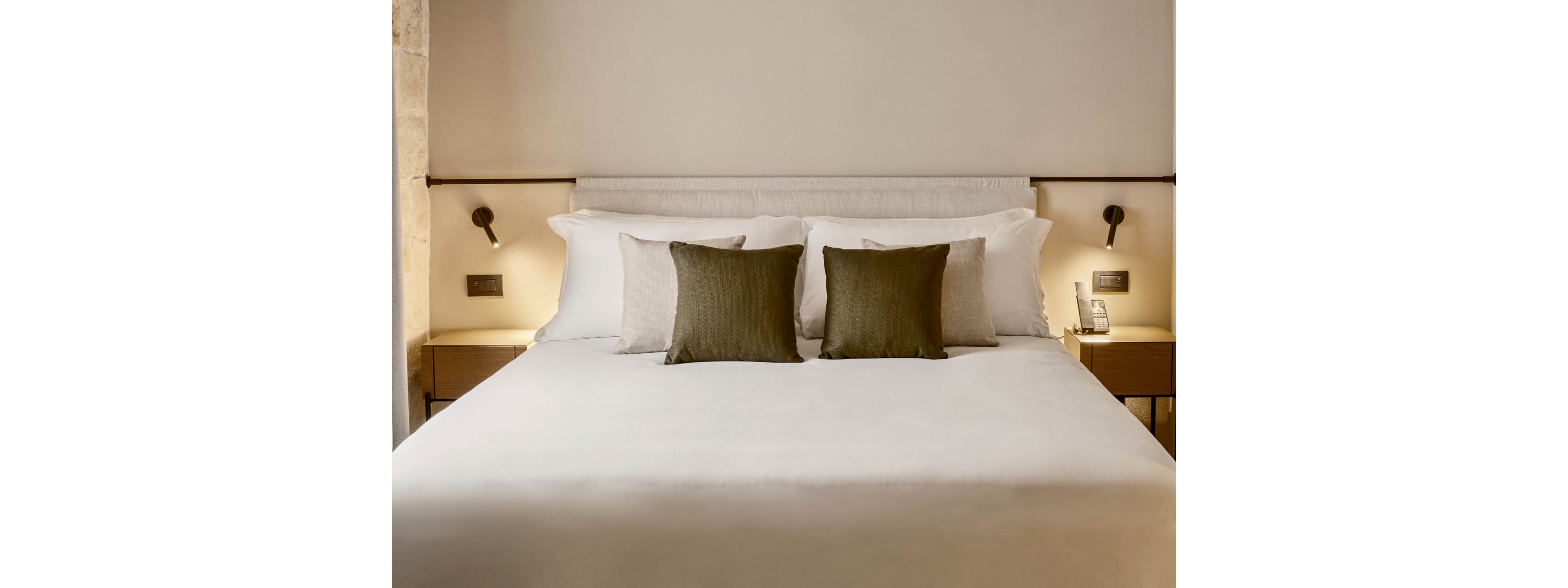A neatly made double bed with crisp white linens and three olive-green accent pillows is centered in a minimalist bedroom. Soft wall-mounted reading lights flank the bed above simple wooden nightstands. The room features neutral tones, clean lines, and a calm, contemporary design. A telephone and small details on the bedside tables suggest a modern hotel setting.