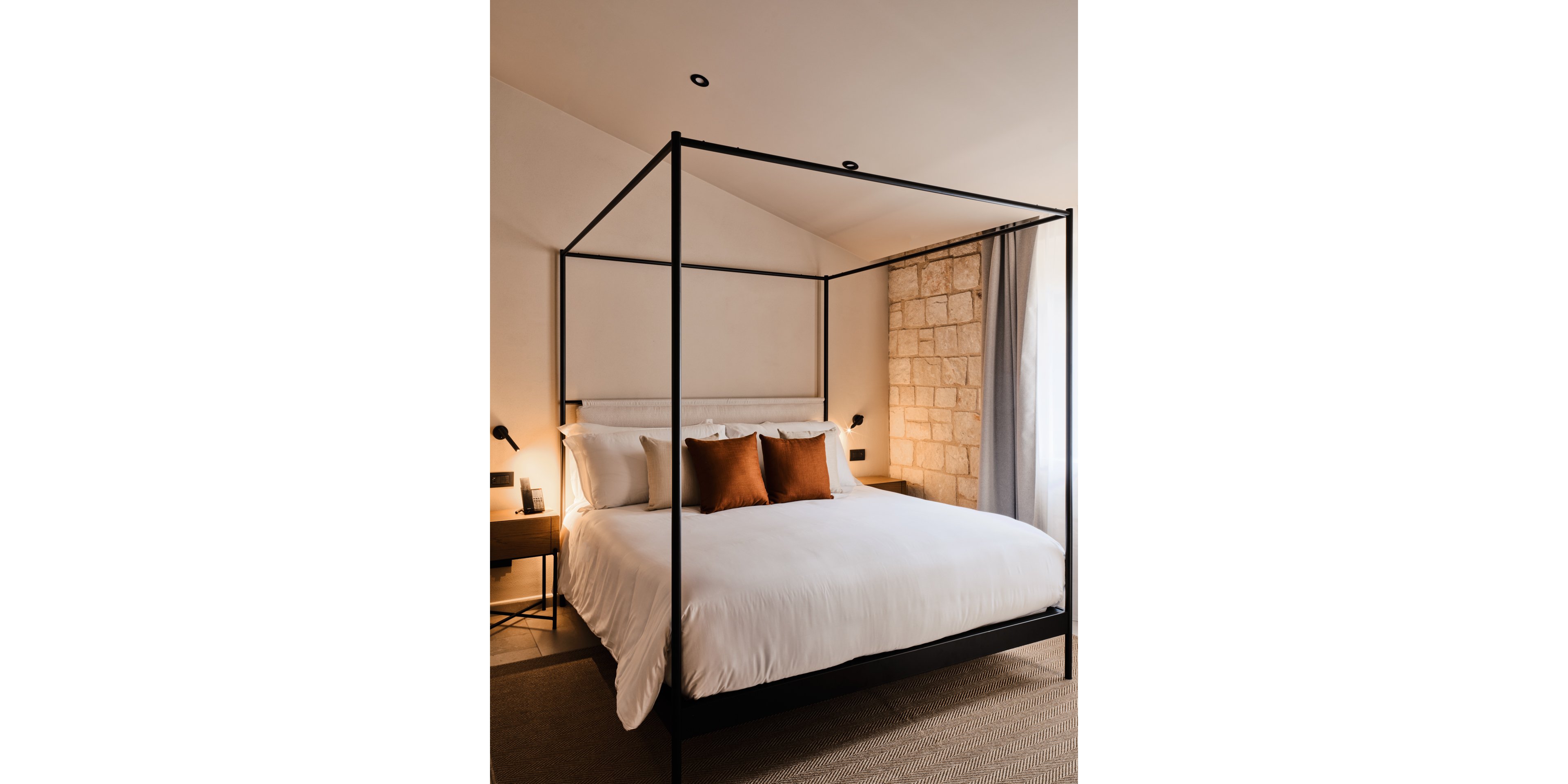 A contemporary four-poster canopy bed with crisp white linens and two brown accent pillows is the focal point of this stylish bedroom. The room features warm lighting, minimalist bedside tables, and wall-mounted reading lamps. A textured stone wall and soft curtains add depth and contrast to the neutral color palette. The overall atmosphere feels inviting, comfortable, and modern.