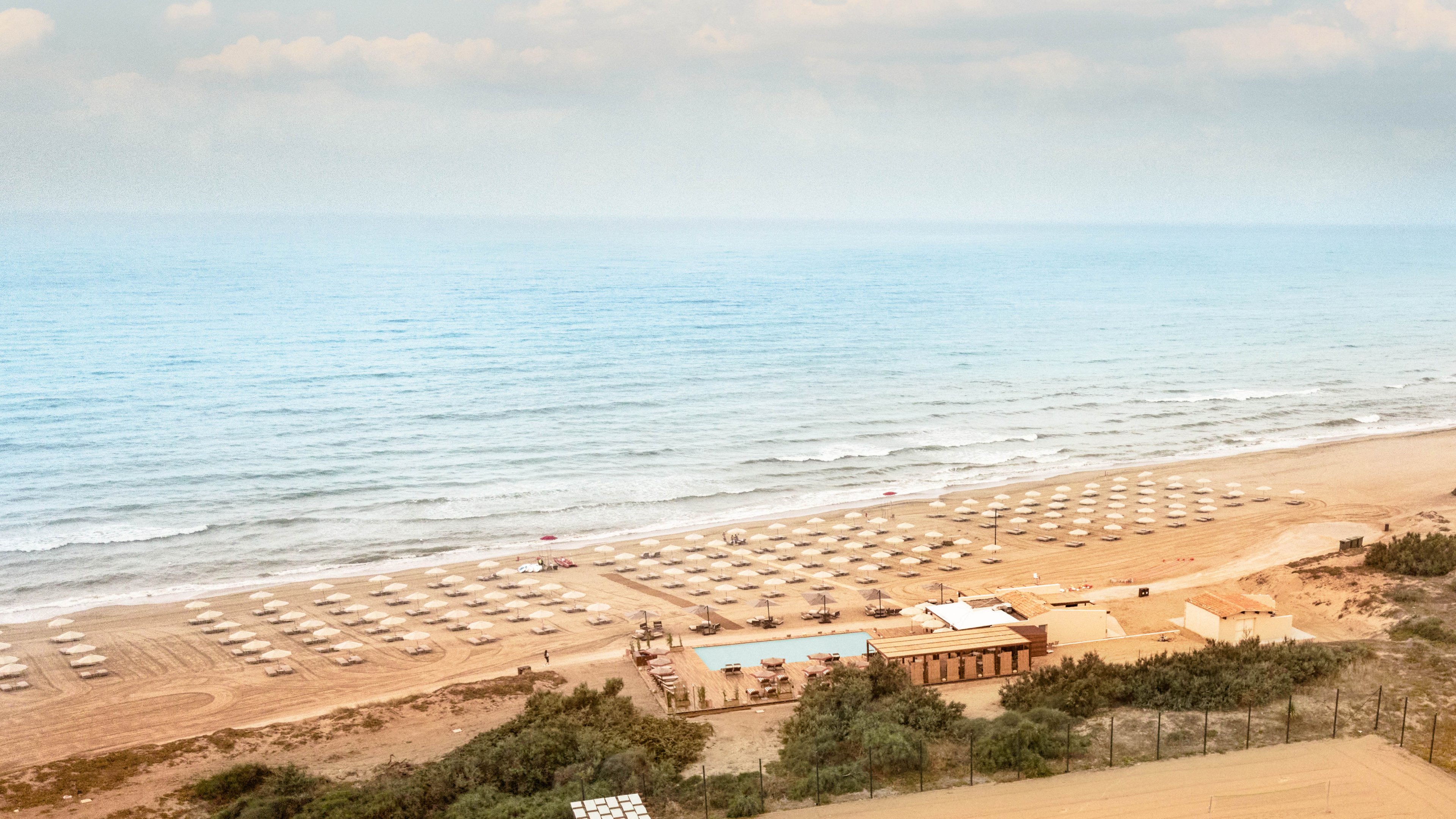 A wide sandy beach stretches along a calm sea, lined with rows of closed white umbrellas and sun loungers. In the middle of the scene, a small resort complex with a swimming pool sits just behind the beach. Gentle waves roll toward the shore under a hazy sky, creating a tranquil coastal atmosphere. Green shrubs and fencing frame the foreground, adding depth to the panoramic view.