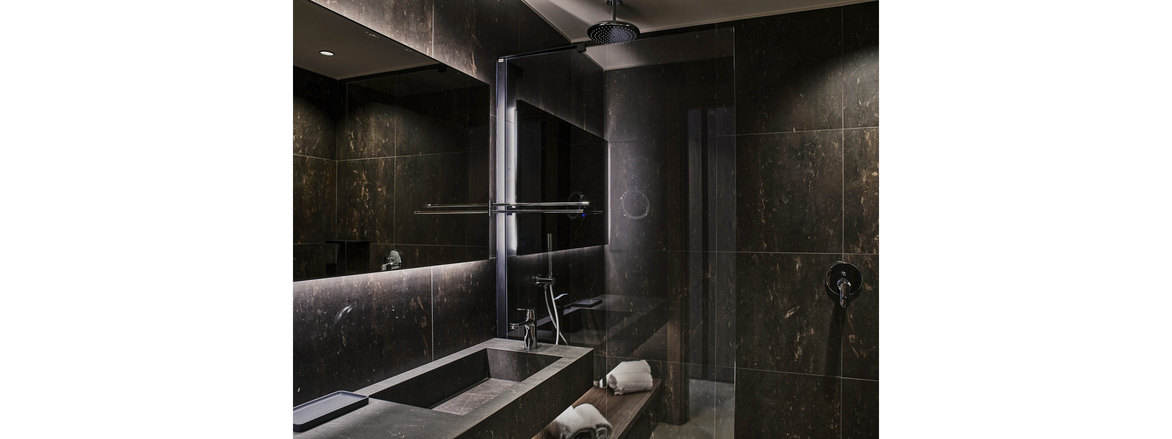 Contemporary bathroom featuring dark stone walls and a sleek rectangular sink. A large mirror with integrated backlighting reflects the walk-in shower area. The shower has a ceiling-mounted rain showerhead and minimalist fixtures. Rolled white towels are neatly placed on a shelf beneath the vanity, enhancing the spa-like atmosphere.