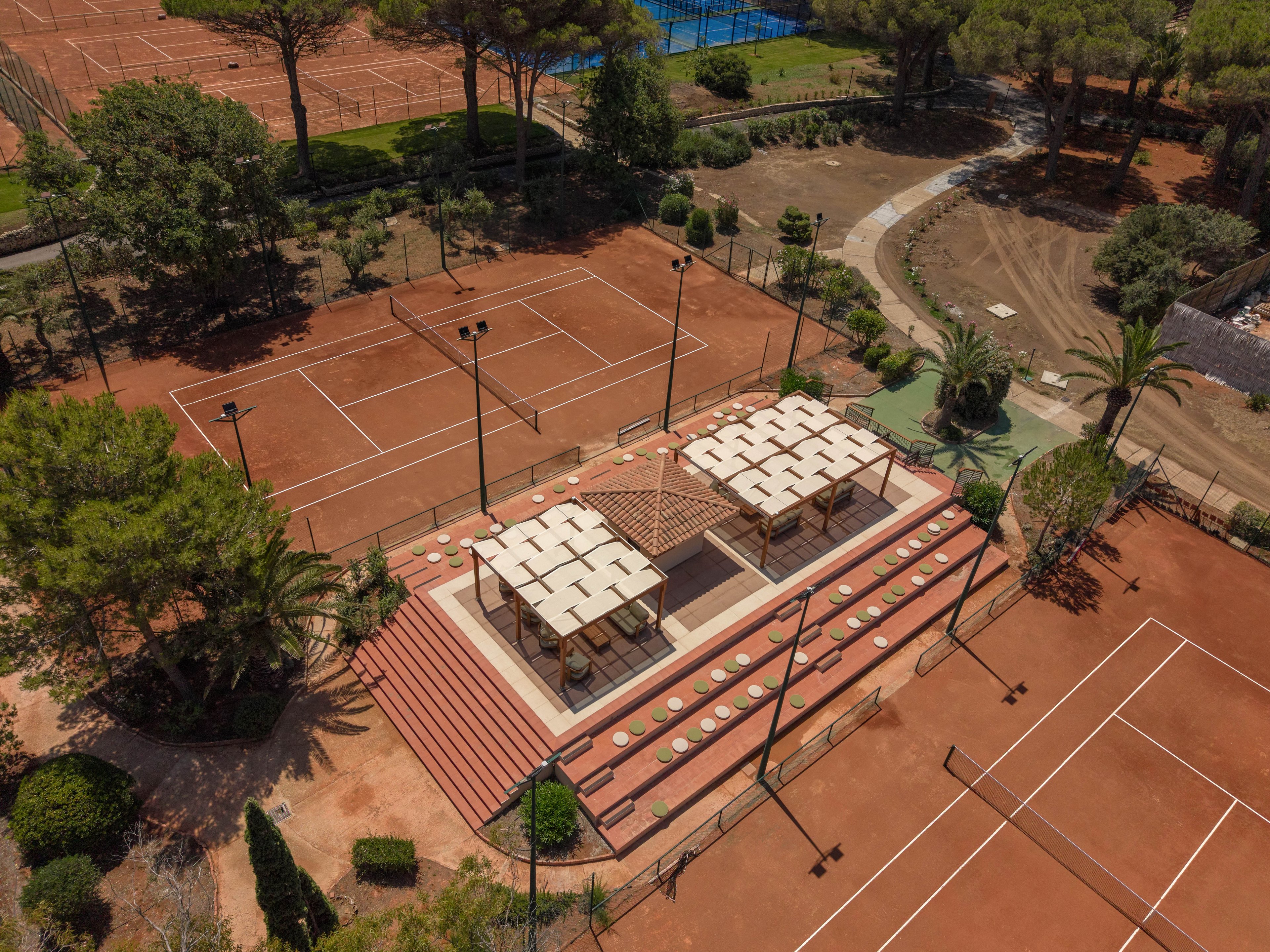 A drone captures an aerial view of a clay tennis court facility surrounded by trees. Multiple red clay courts extend around a central pavilion with shaded seating and steps. Tall light poles line the courts, and pathways and landscaped areas frame the sports complex. The scene suggests a quiet, sunny day at an outdoor tennis club.