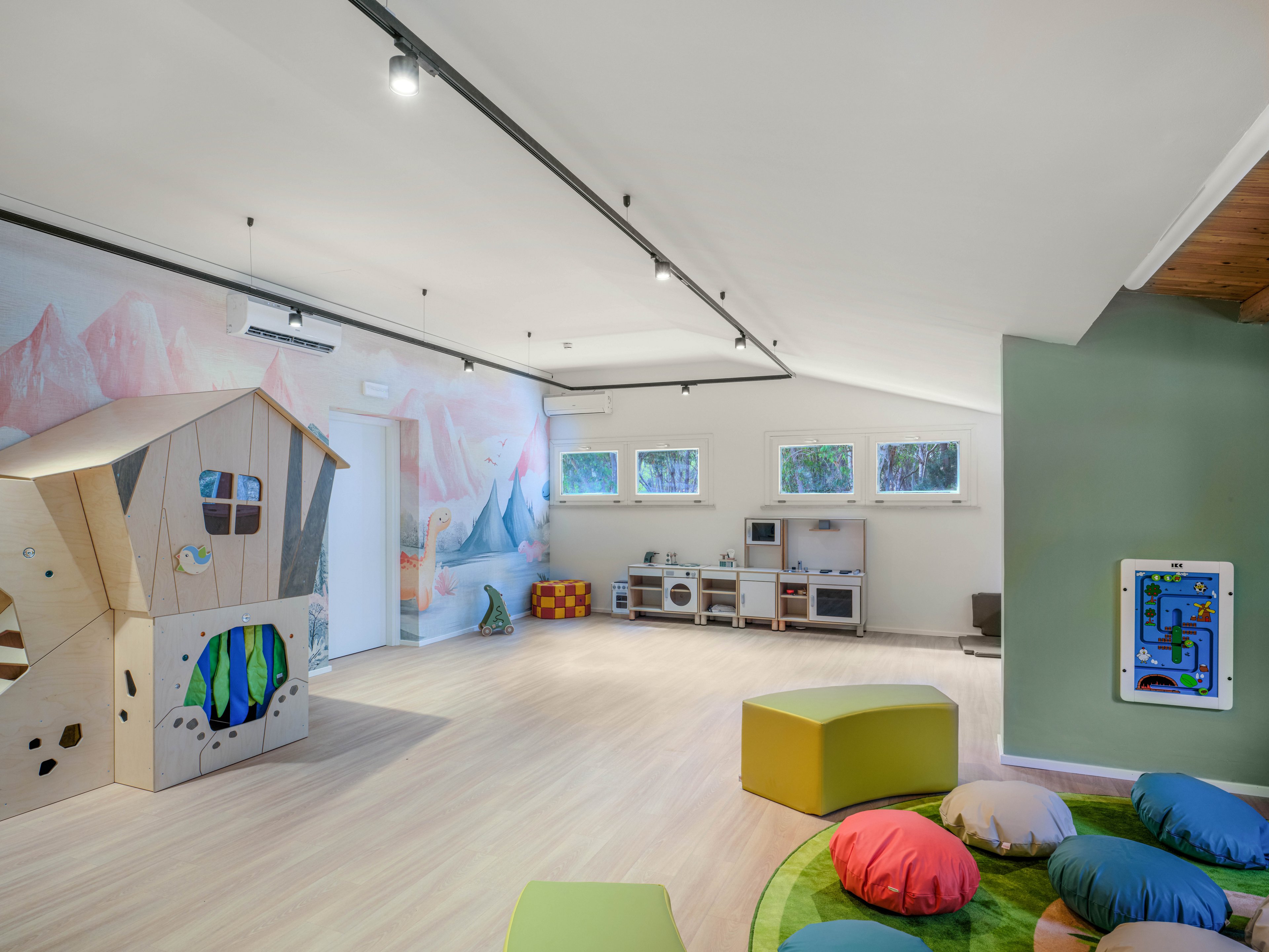 Spacious children’s playroom featuring colorful soft seating and a wooden playhouse structure. The room includes a play kitchen area with toy appliances along one wall and a wall-mounted activity board. Large windows bring in natural light, highlighting pastel wall murals and a green circular rug with cushions. The overall atmosphere is clean, safe, and inviting for kids’ activities and learning.