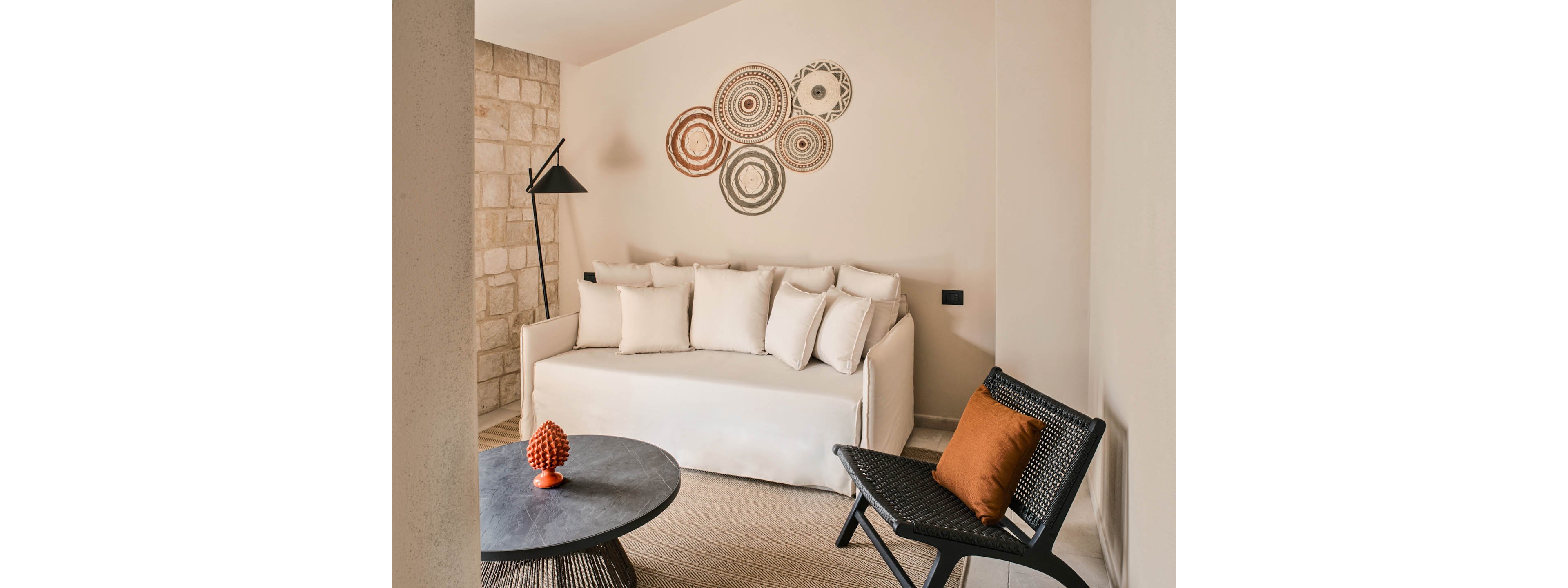 A minimalist living room features a white sofa filled with plush cushions against a light beige wall. Circular decorative wall art adds visual interest above the sofa, while a black floor lamp stands to the side. In the foreground, a round dark coffee table holds an orange sculptural object, and a woven black chair with an orange cushion complements the palette. The space combines soft neutral tones with warm accents for a contemporary, inviting atmosphere.
