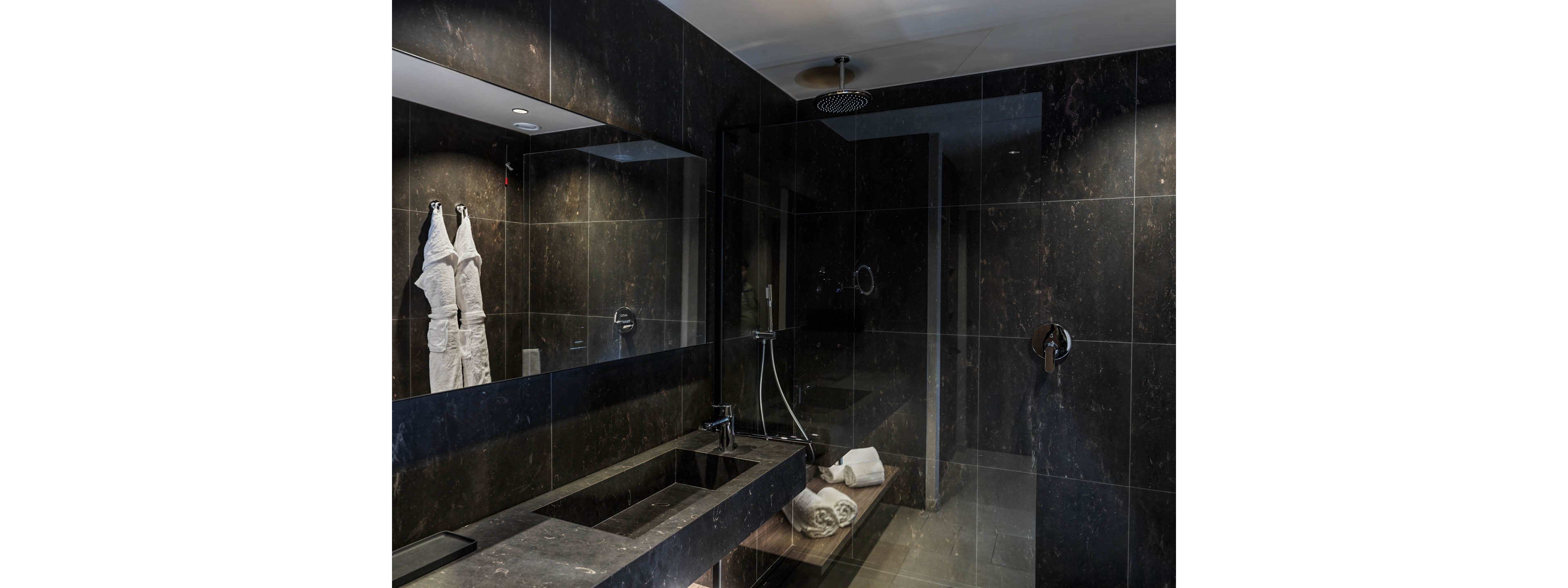 Contemporary bathroom featuring dark marble walls and countertop with a sleek rectangular sink. A glass-enclosed walk-in shower with a ceiling-mounted rain showerhead occupies the back of the room. White bathrobes hang neatly on the wall and folded towels rest on a bench inside the shower area. Subtle recessed lighting and reflective surfaces create a sophisticated, spa-like atmosphere.