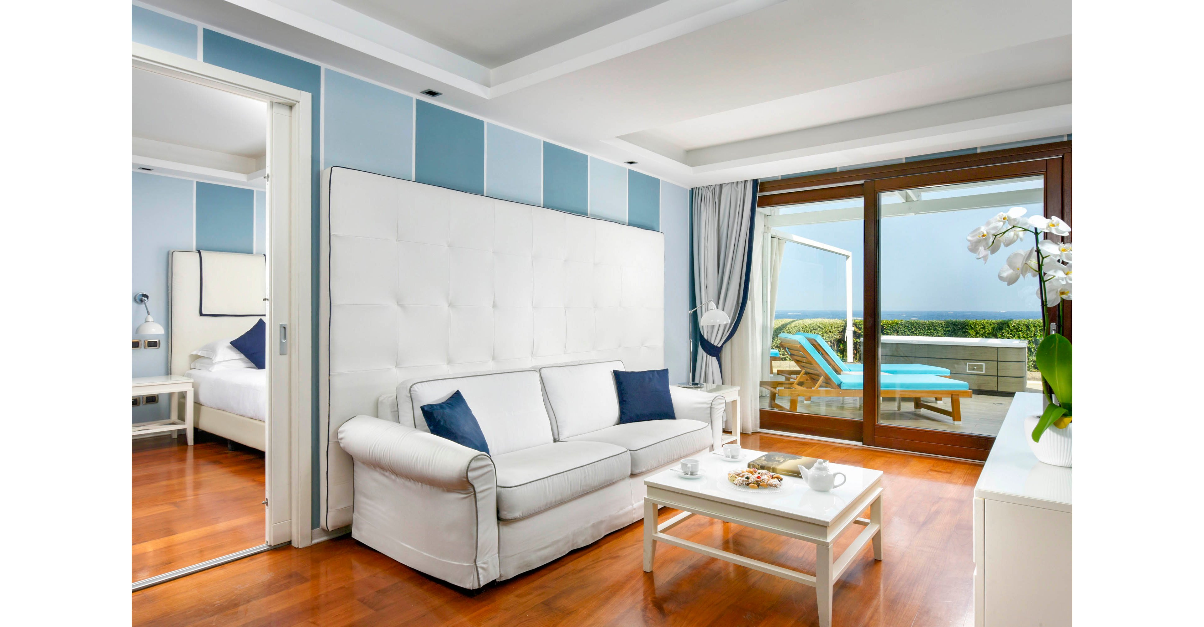A modern hotel suite features a white sofa with navy accent pillows against a tall upholstered headboard wall. The room opens onto a terrace with sun loungers and a view of the ocean beyond large sliding glass doors. Light blue wall panels, white curtains, and polished wooden floors create a fresh coastal atmosphere. A coffee table with cups, saucers, and snacks adds a welcoming touch to the bright interior.