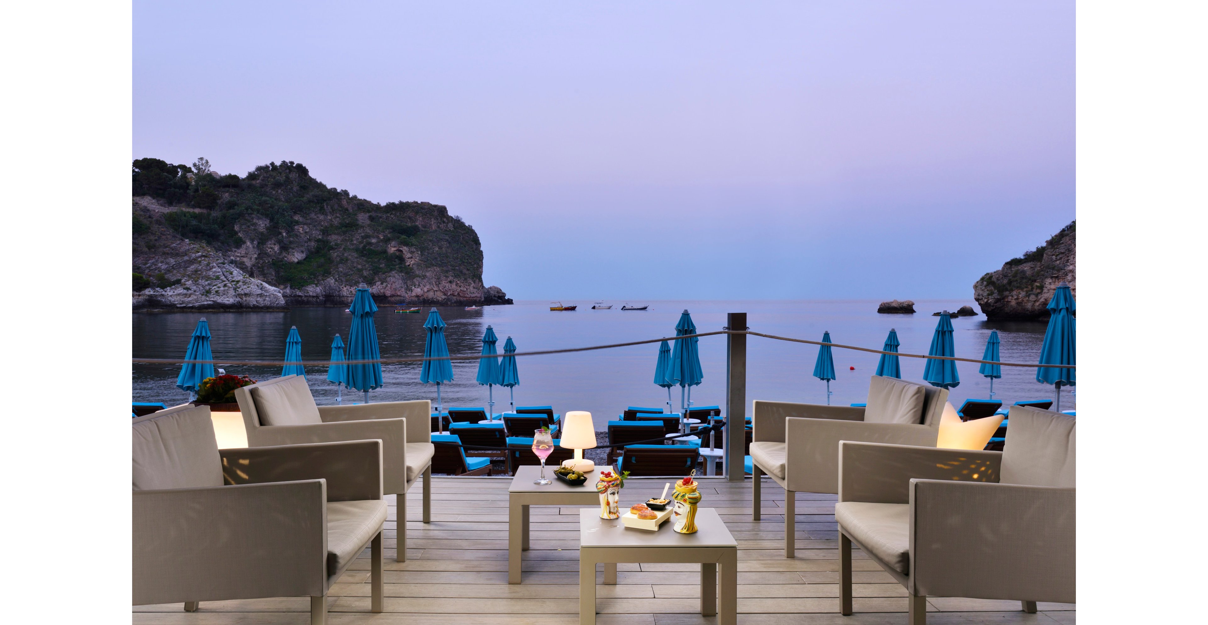 A modern outdoor lounge area overlooks a tranquil bay with rocky cliffs and calm waters at dusk. Four white armchairs surround two tables set with cocktails, wine, and plated appetizers. Blue umbrellas and lounge chairs line the waterfront, creating a serene and inviting atmosphere for relaxation by the sea.