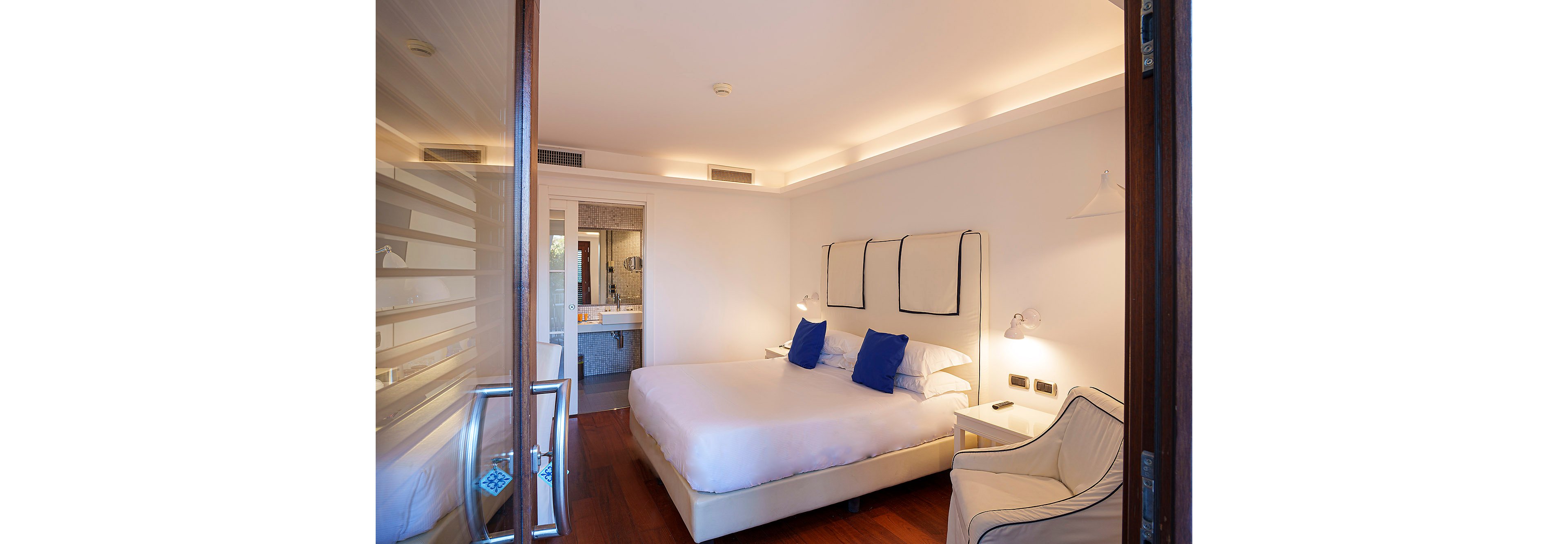 A contemporary hotel room features a large bed with crisp white linens and two blue accent pillows. The space is illuminated by soft, indirect lighting and includes a white armchair and bedside lamps. The room has wooden flooring and an open view into a sleek bathroom. The overall mood is calm and inviting, with a minimalist style.