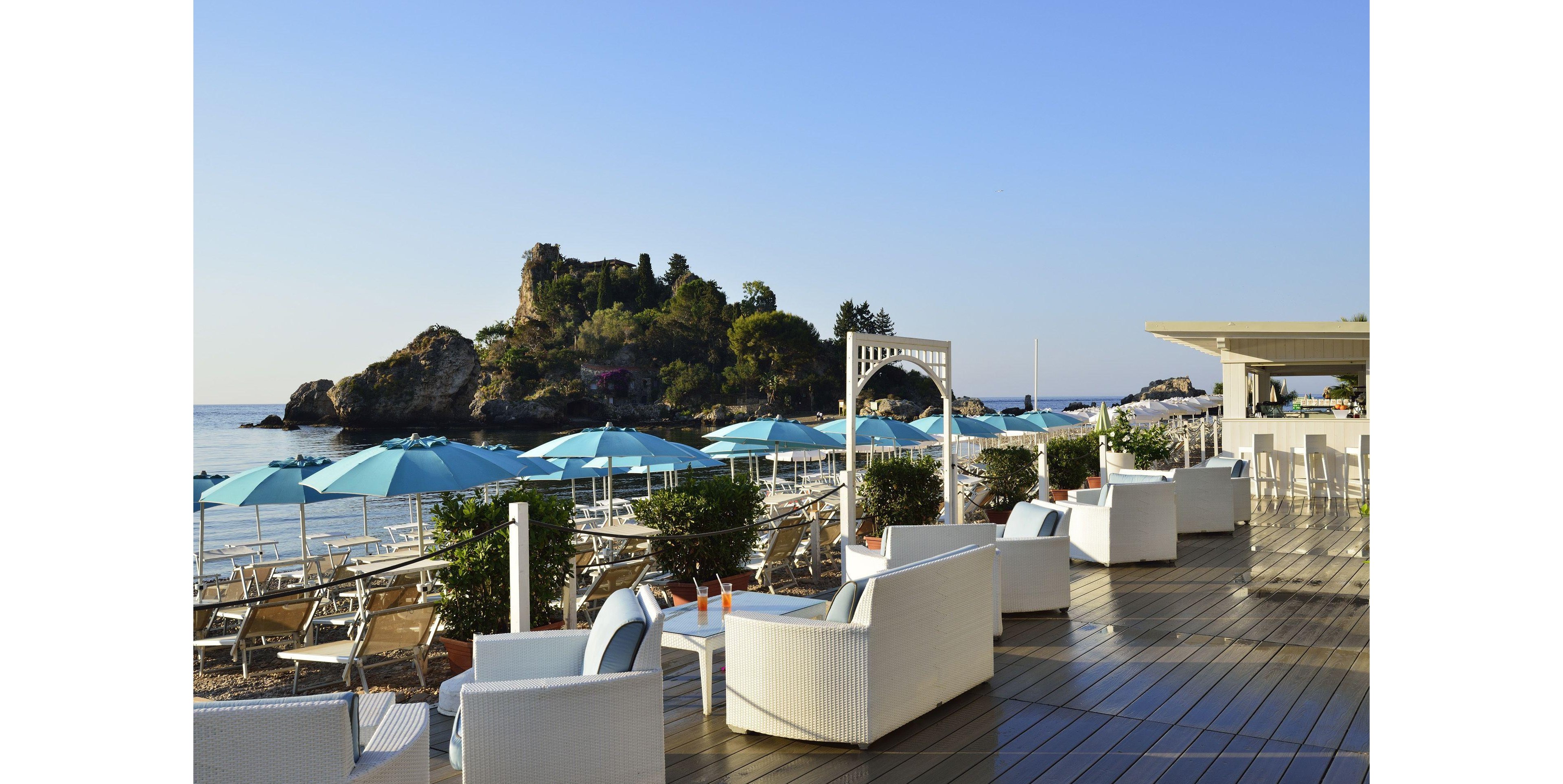 A stylish outdoor lounge area overlooks a scenic coastline with a small rocky island in the background. The setting features white wicker seating, turquoise umbrellas, and a wooden deck, creating a relaxed and inviting atmosphere. The clear sky and calm sea enhance the tranquil mood, while drinks on the tables suggest leisure and refreshment. No visible text or numbers are present in the image.