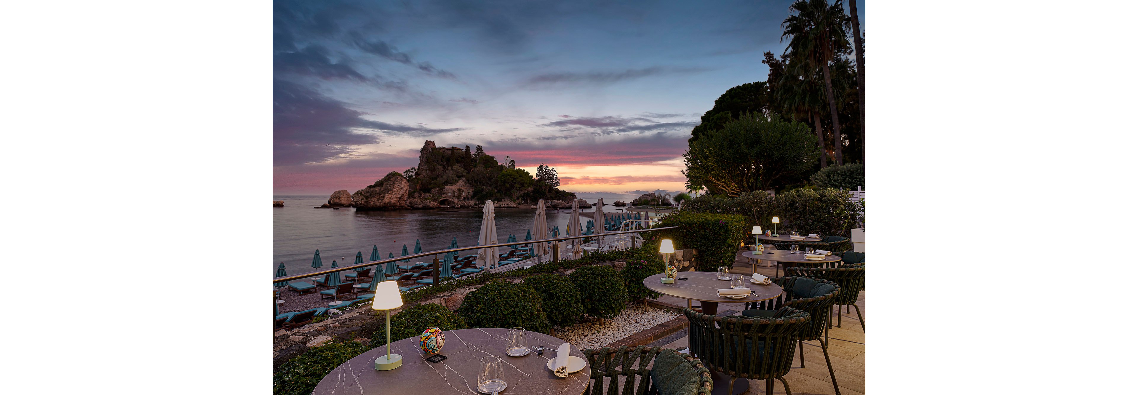An elegant outdoor restaurant terrace overlooks a tranquil bay at sunset, with small tables set for dining. The scene features closed umbrellas and lounge chairs along the beach, with a rocky island and calm water in the background. Soft lighting from table lamps creates a cozy atmosphere, while lush greenery frames the seating area. The sky displays a gradient of blue, pink, and orange hues, enhancing the serene mood.