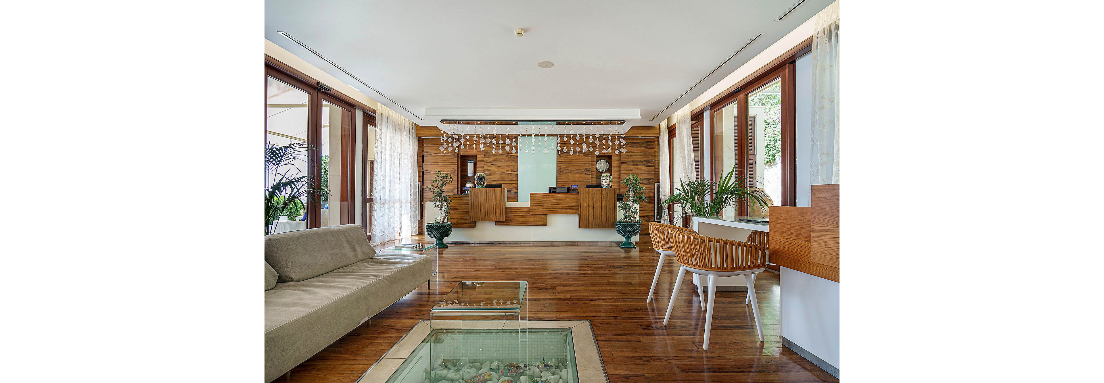 A contemporary hotel reception area features a sleek wooden front desk and polished hardwood floors. The space is decorated with potted plants, modern furniture, and large windows allowing natural light to fill the room. Decorative hanging elements and a glass-topped table with seashells add visual interest. The overall mood is welcoming and tranquil, with a neutral color palette and minimalist style.