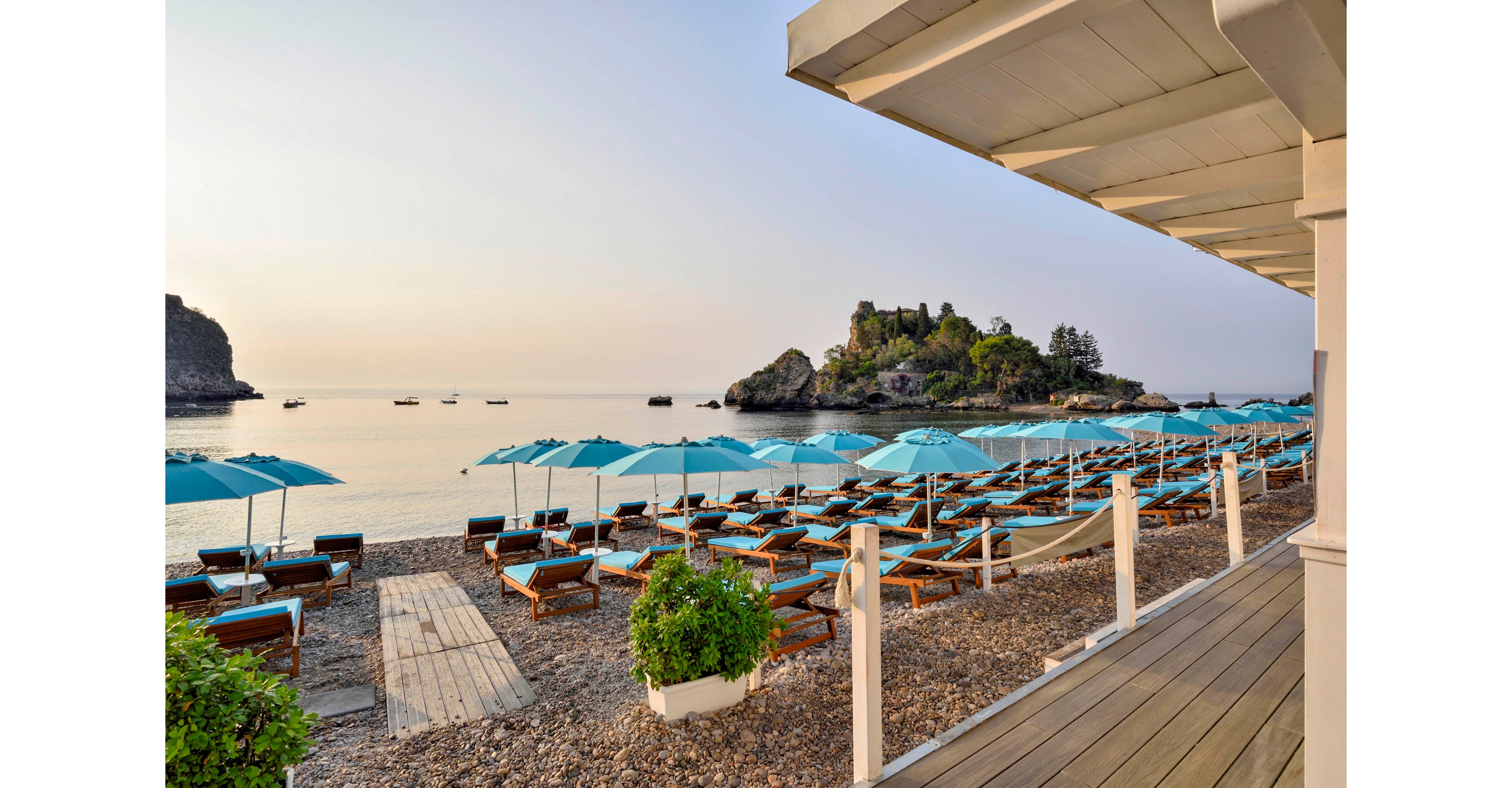 A tranquil beach resort features rows of turquoise umbrellas and wooden lounge chairs arranged on a pebbled shore. The calm sea stretches out to small boats and a rocky island in the distance, while the foreground includes a shaded deck area with potted plants. The scene is bathed in soft, early morning or late afternoon light, creating a peaceful and inviting atmosphere.