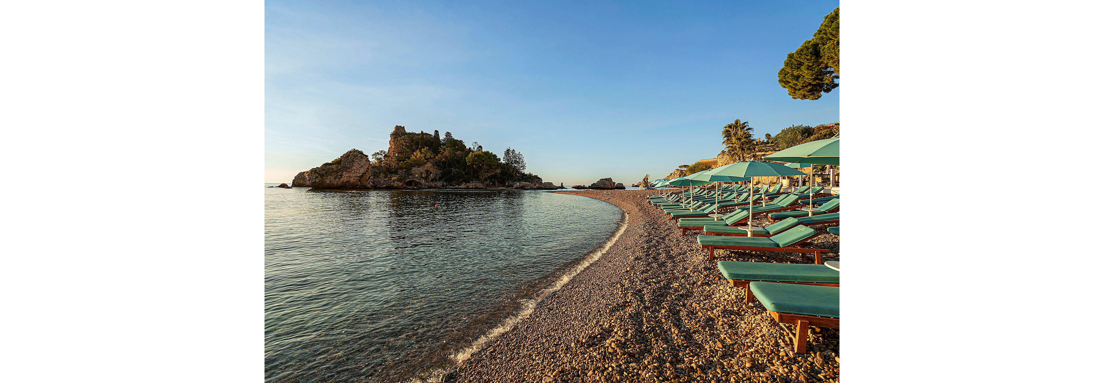 A tranquil pebble beach curves along calm, clear water under a golden sunrise. Rows of turquoise sun loungers and matching umbrellas line the shore, all unoccupied, creating a peaceful and inviting atmosphere. In the distance, a small rocky island and lush greenery add to the scenic coastal setting.