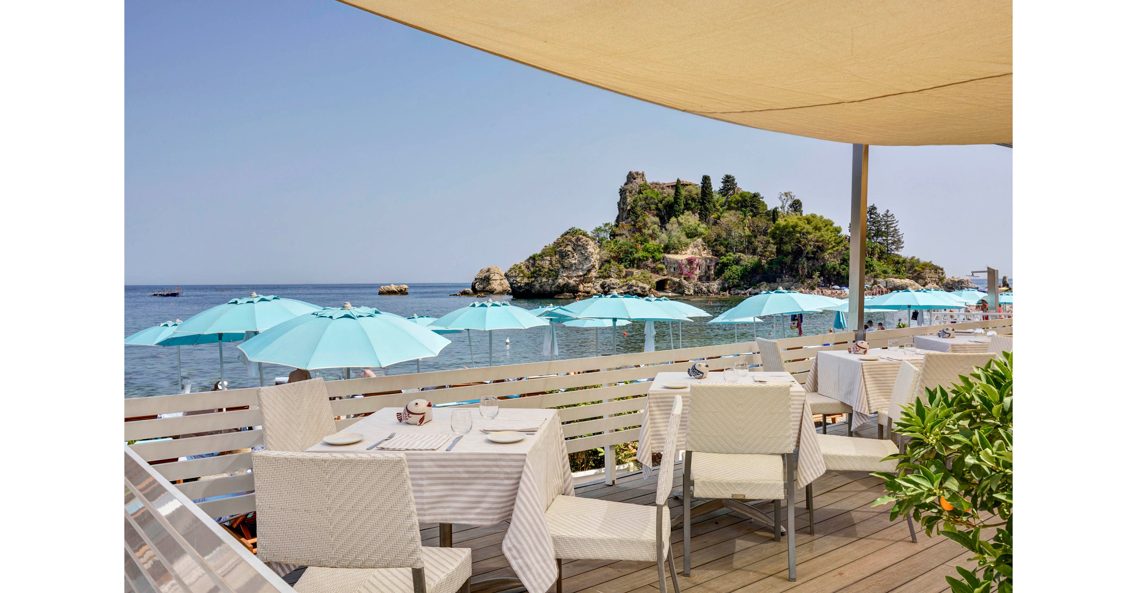 Outdoor dining tables are set on a wooden terrace overlooking a scenic beach. Turquoise umbrellas line the shore, providing shade for beachgoers, while a rocky island rises in the background. The setting is bright and tranquil, with neutral table linens and a clear blue sky creating a relaxed atmosphere. No visible text or numeric values are present in the image.