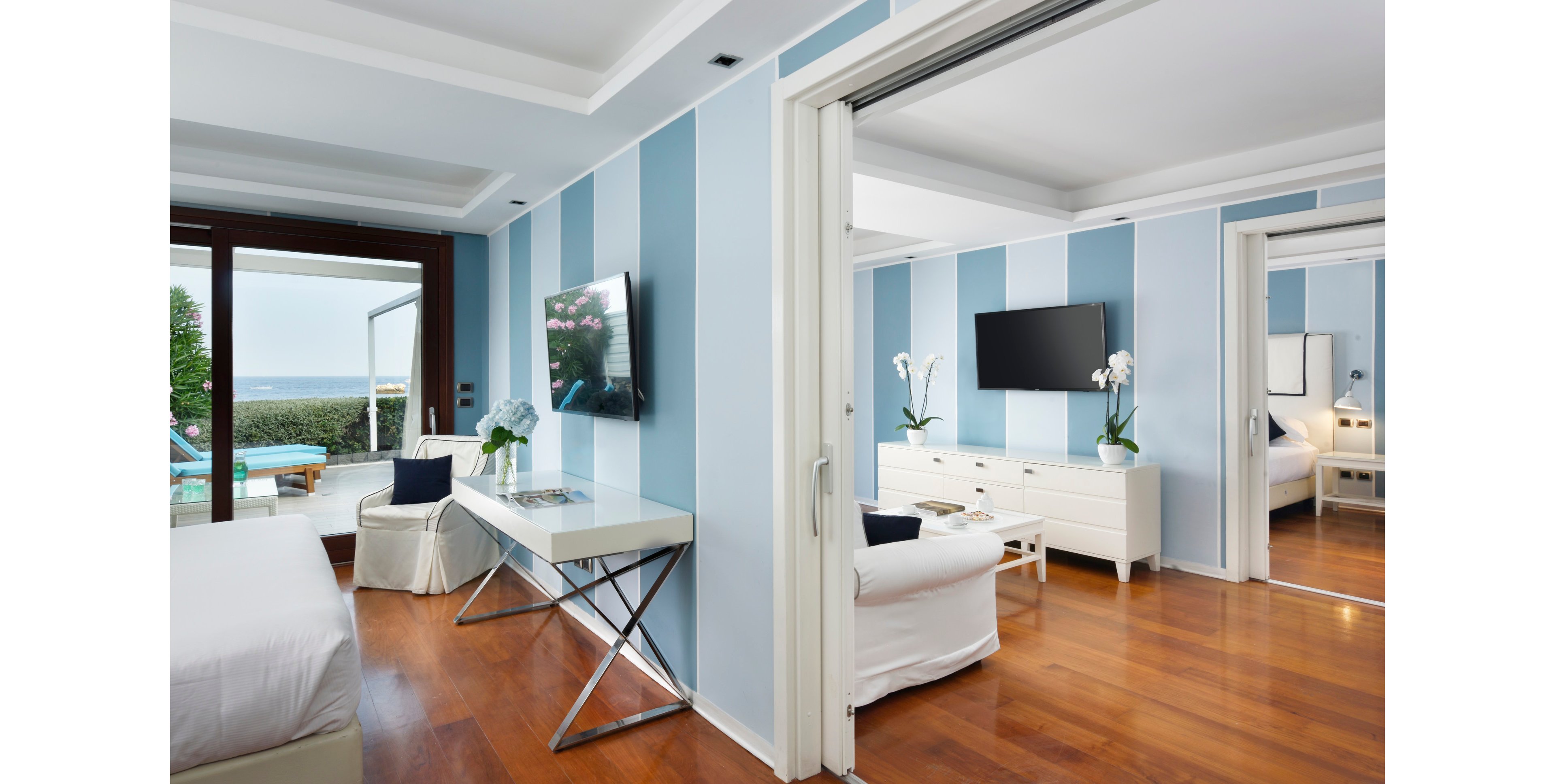 A contemporary hotel suite features blue and white striped walls, hardwood floors, and sleek white furniture. The room is divided into two spaces, each with a flat-screen television and large windows offering a scenic ocean view. Decorative orchids and minimalist decor create a fresh, relaxing atmosphere, with natural light enhancing the coastal vibe.